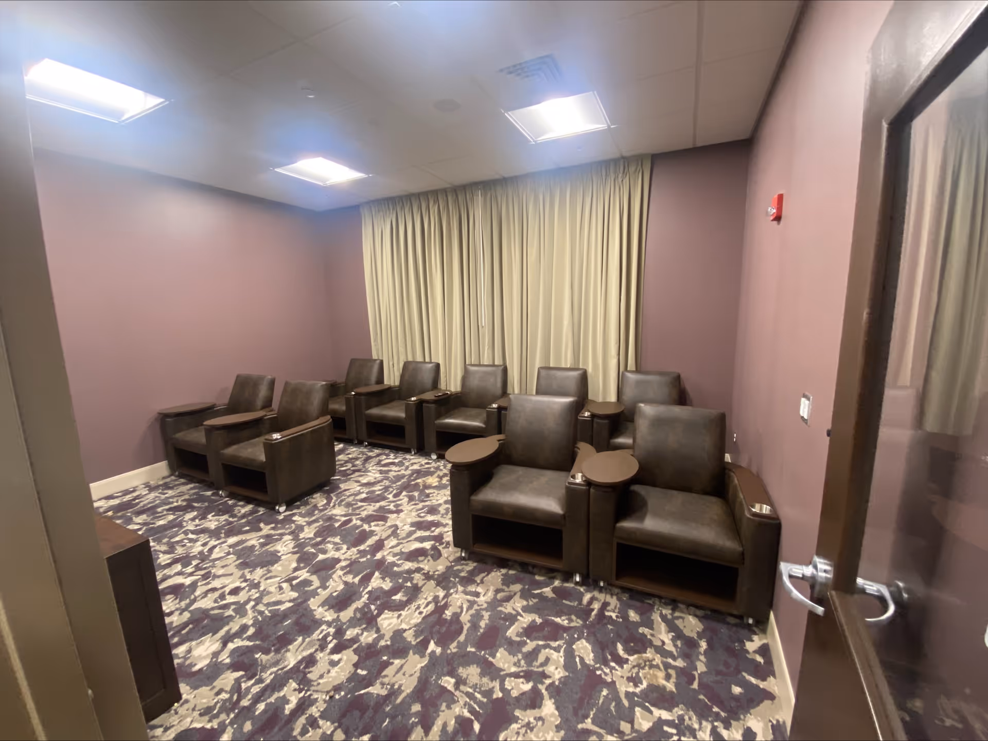 A small room with eight brown leather armchairs arranged in two rows facing a blank wall. Each chair has a small attached wooden table. The room has purple walls, a patterned carpet in shades of purple and beige, and a large beige curtain covering the window. The ceiling has recessed lighting panels, and there is a glass door partially open on the right side.