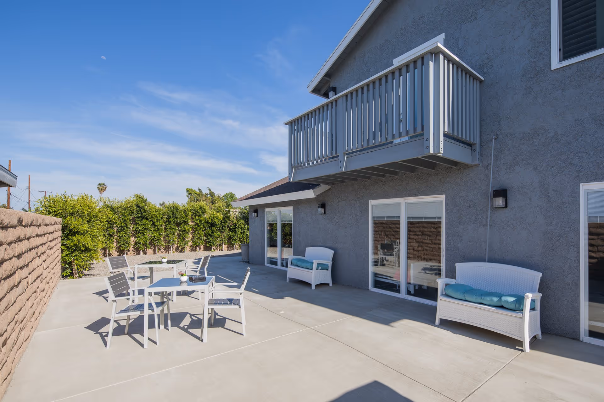 Sunlit outdoor patio with white patio furniture, a balcony above, and sliding glass doors on a gray stucco building.