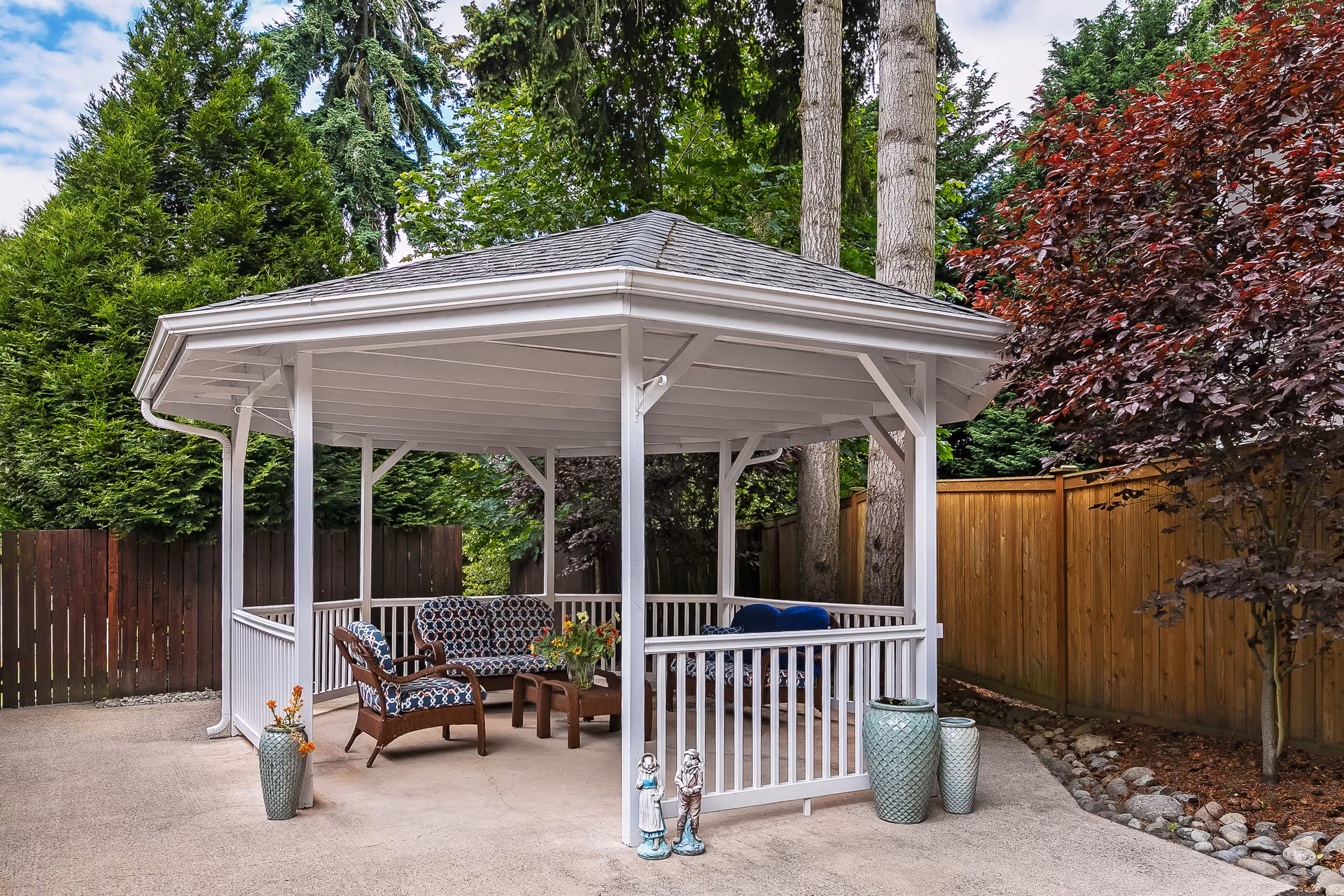 A white wooden gazebo with a gray roof situated on a concrete patio surrounded by a wooden fence and lush green trees. Inside the gazebo, there are cushioned chairs and a small table with a flower arrangement. Decorative vases and small statues are placed around the gazebo.