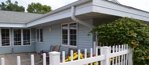 Blue single-story senior living building exterior with white picket fence, patio, and flowers.