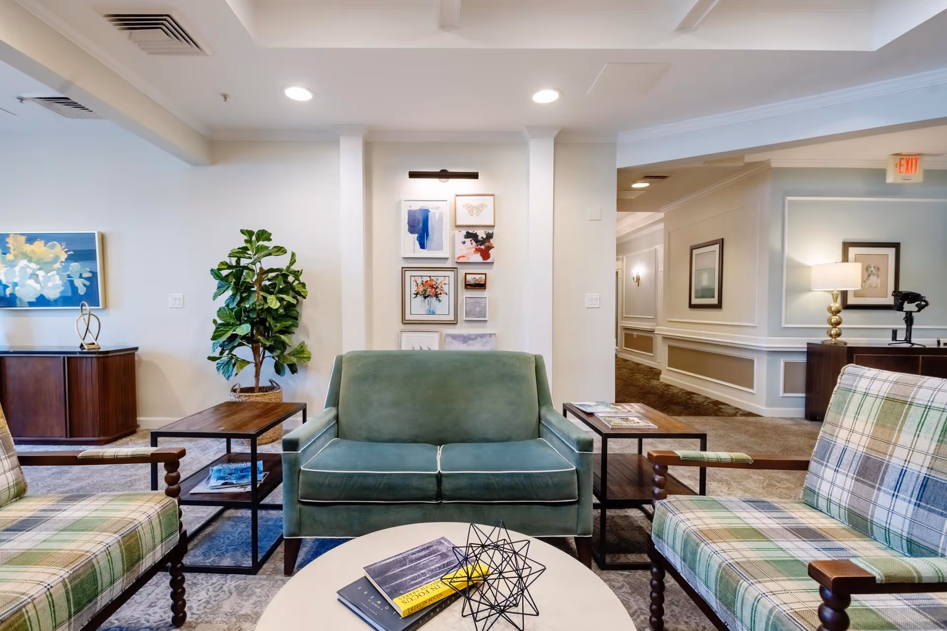 A cozy living room area in Blue Bell Place featuring a green loveseat centered between two plaid armchairs. There are two wooden side tables with magazines, a round white coffee table with books and a decorative object, a potted plant, framed artwork on the wall, and soft lighting creating a welcoming atmosphere.