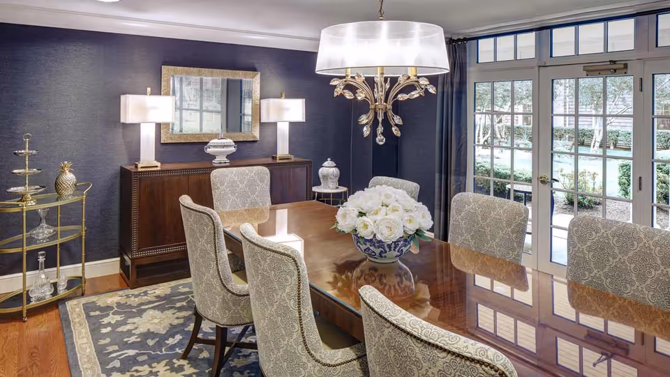 Elegant dining room with a polished wooden table surrounded by eight patterned upholstered chairs. A large floral centerpiece sits on the table. The room features a dark blue textured wall with a wooden sideboard, two table lamps, and a decorative mirror. A chandelier with crystal accents hangs above the table. French doors with glass panes open to a garden outside. A patterned area rug covers the hardwood floor.