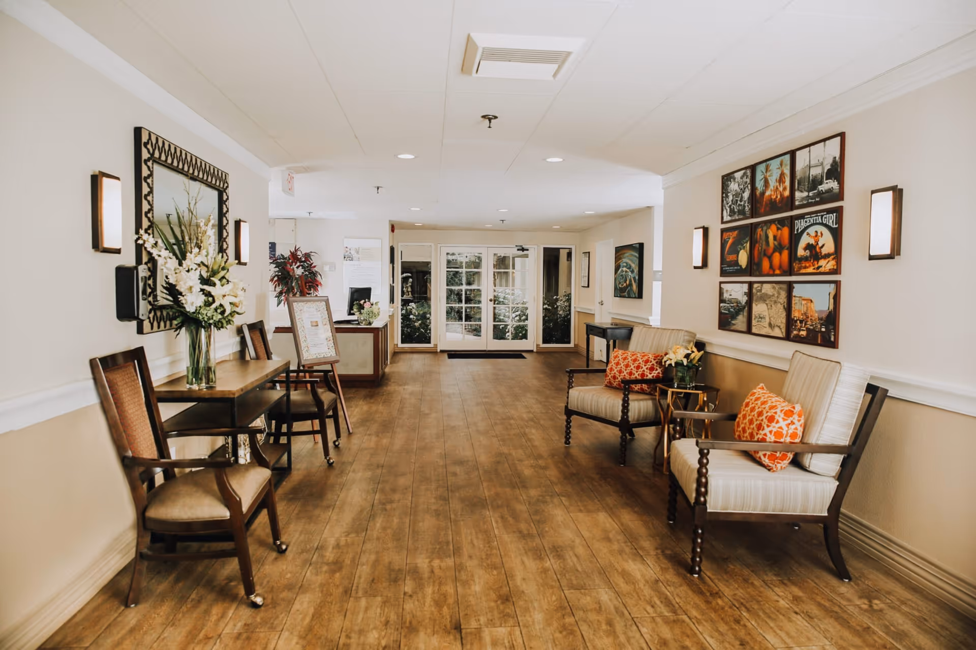 A well-lit hallway in a senior living facility with wooden flooring, decorated with chairs on both sides. On the left, there is a table with a vase of white flowers and a large mirror above it. On the right, two cushioned chairs with orange patterned pillows are placed next to a small side table with a flower arrangement. The walls have framed pictures and wall-mounted lights. At the end of the hallway, glass double doors lead outside to a garden area.