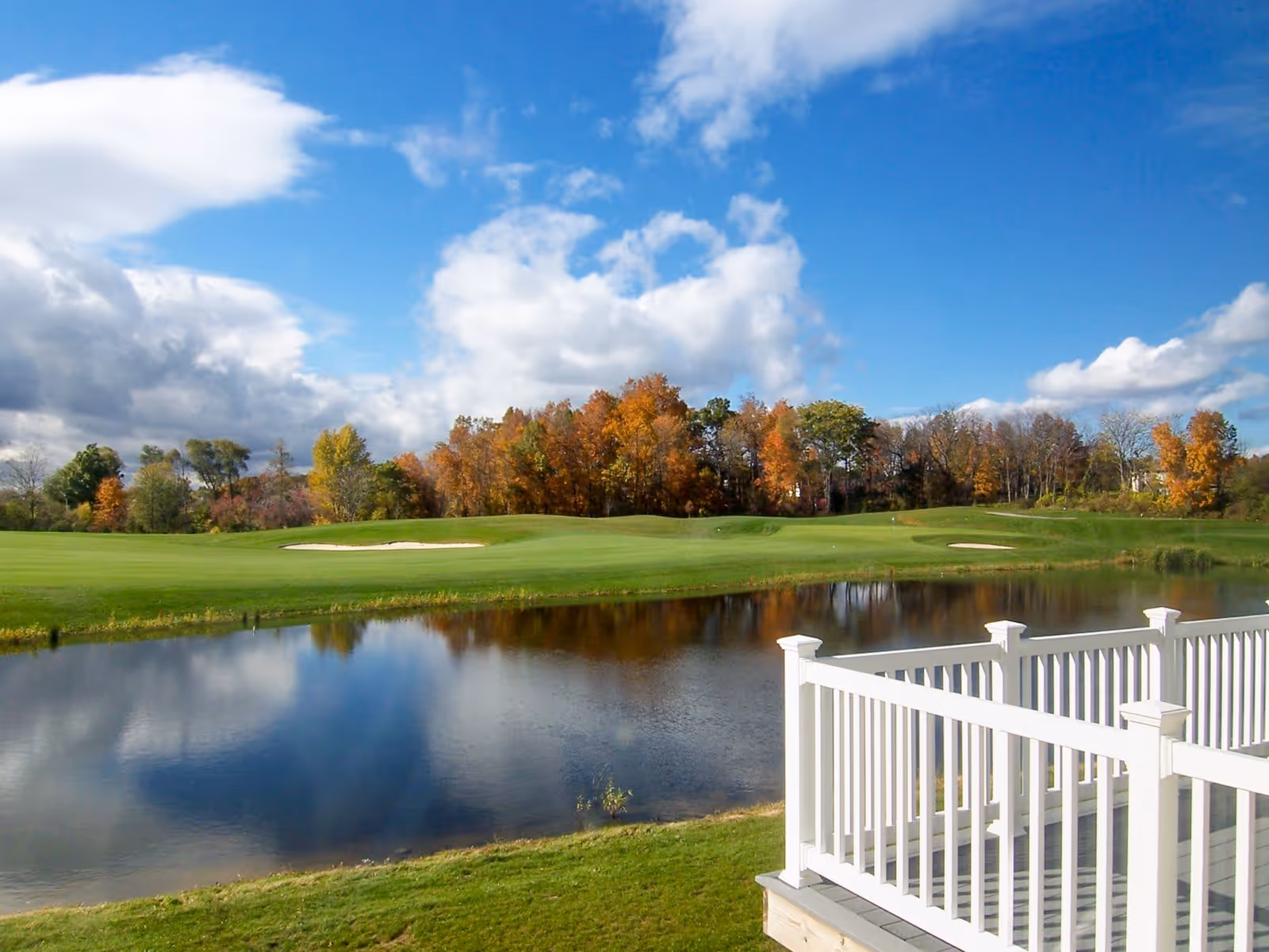 View of a golf course with a pond in the foreground and a white railing of a deck on the right side. Trees with autumn foliage are visible in the background under a partly cloudy blue sky.