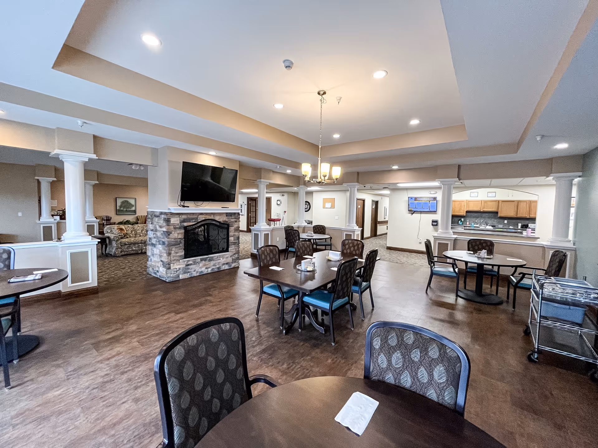 A spacious dining area in a senior living facility with several round and rectangular tables surrounded by chairs. The room features a stone fireplace with a mounted flat-screen TV above it, white columns, and a chandelier hanging from the ceiling. In the background, there is a kitchen area with wooden cabinets and a TV mounted on the wall.