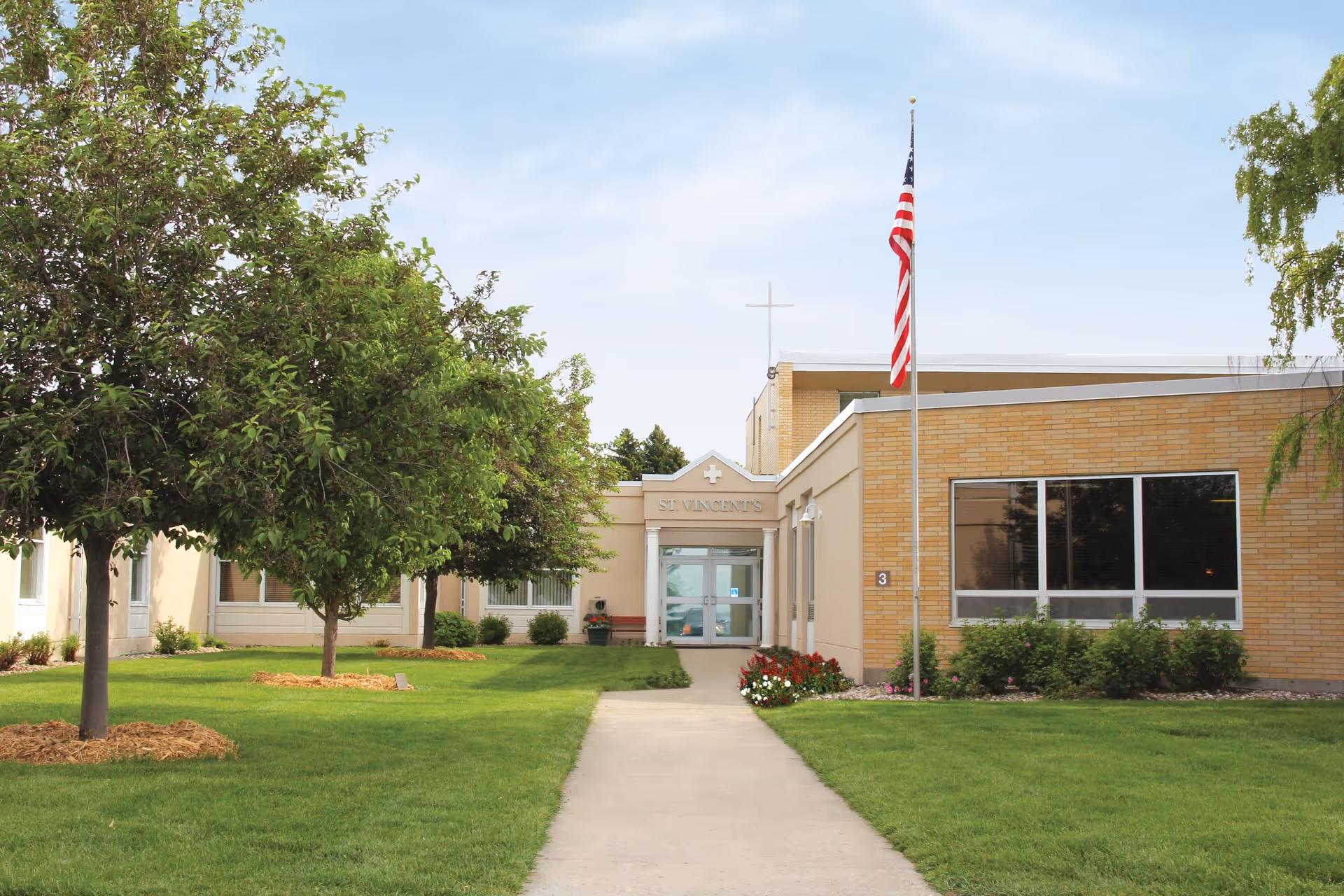 Front entrance of Good Samaritan Society – St. Vincent's with a walkway, lawn, trees, and an American flag on a flagpole.