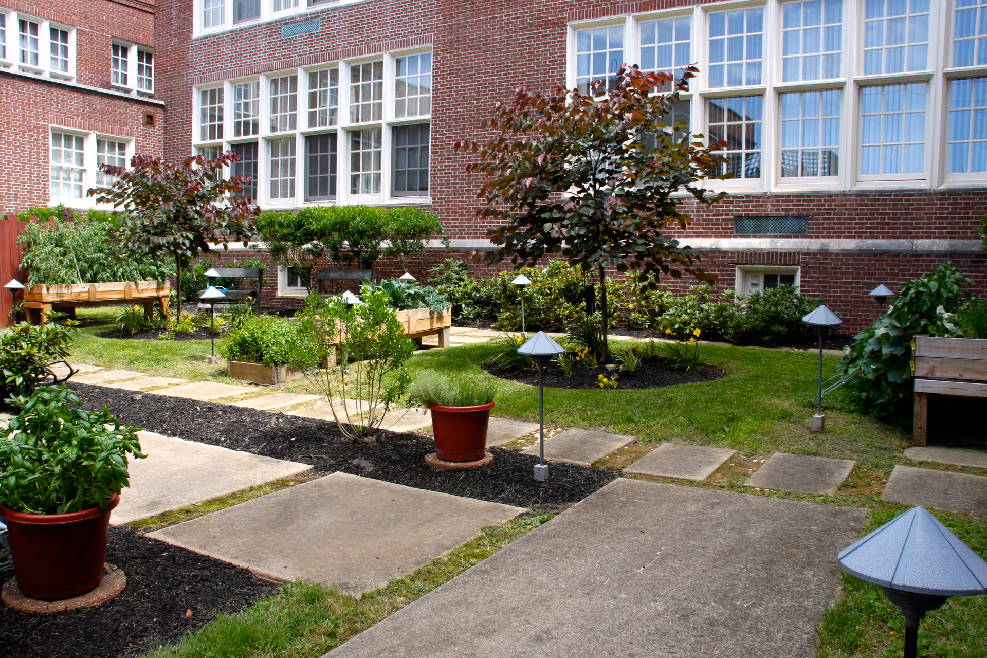 A well-maintained outdoor garden area at The Lorelton with paved walkways, potted plants, small trees, raised garden beds, and decorative garden lights. The garden is adjacent to a brick building with large windows.