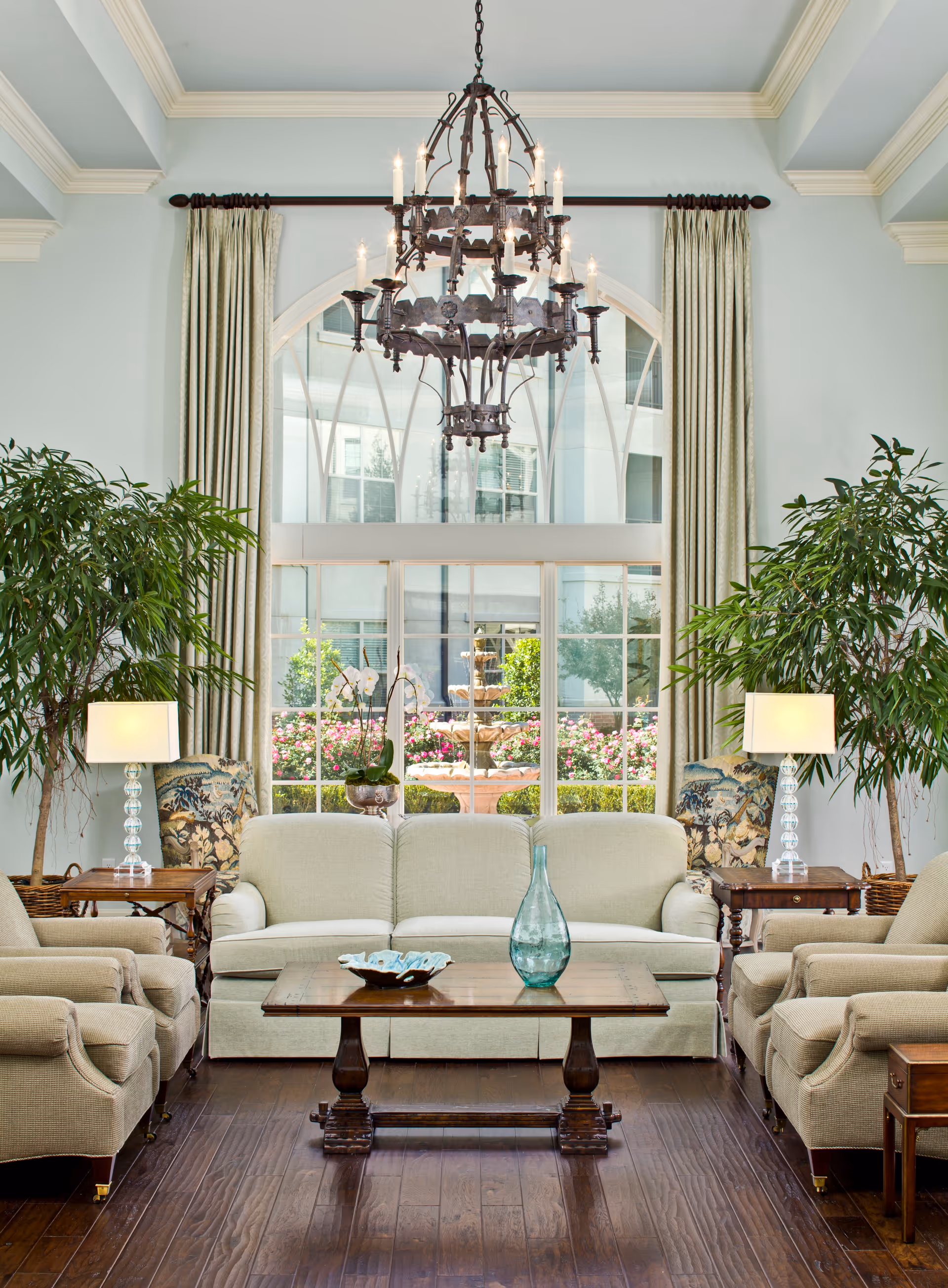 A bright and elegant living room with a large window showing a garden with a fountain outside. The room features a beige sofa and two matching armchairs around a wooden coffee table with decorative items. Two side tables with lamps and large green plants flank the seating area. A large ornate chandelier hangs from the ceiling.