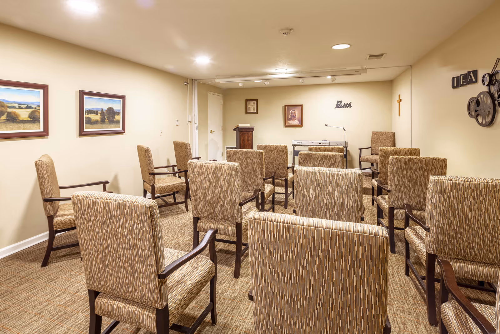 A small meeting or chapel room with several upholstered chairs arranged in rows facing a podium and an electronic keyboard. The walls are decorated with framed pictures, a cross, and the word 'Faith'. The room has beige walls and carpeted floor with overhead lighting.