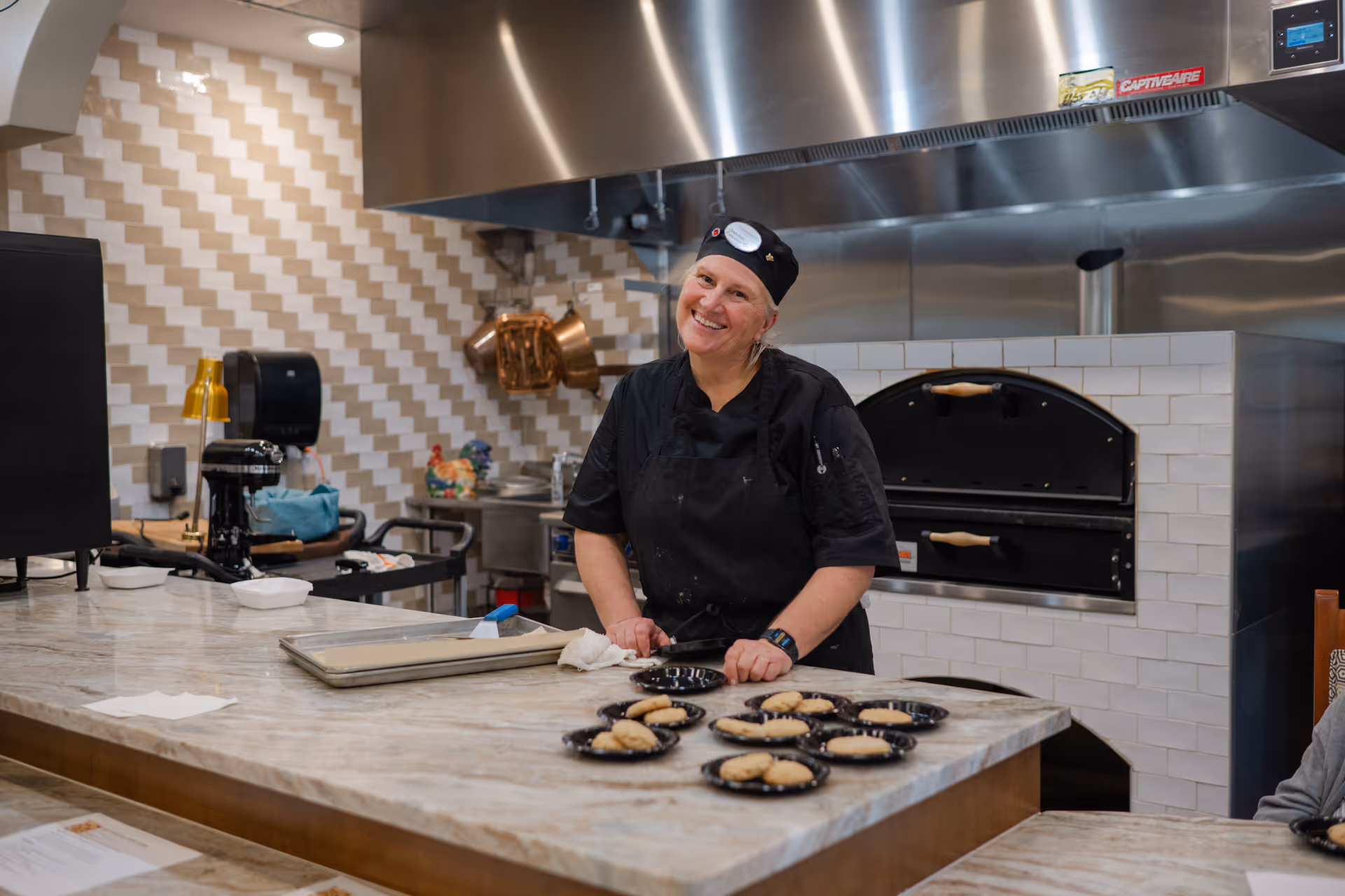 A smiling chef wearing a black uniform and hat stands behind a marble countertop in a kitchen. Several plates with cookies are arranged on the counter. The kitchen features a tiled wall with a geometric pattern, copper pots hanging, and a large stainless steel oven in the background.