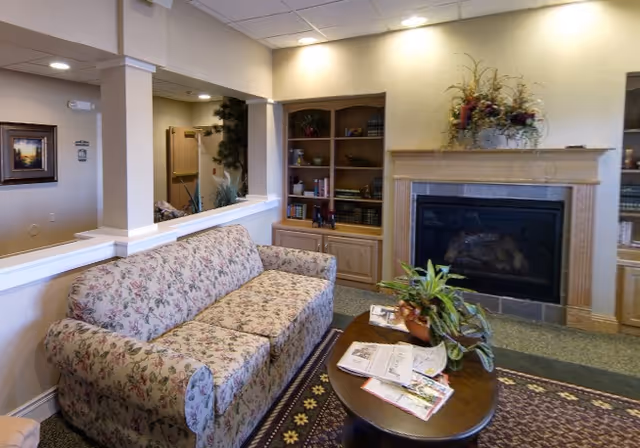 A cozy living room area with a floral patterned sofa, a wooden coffee table with plants and newspapers on it, a fireplace with a decorative floral arrangement on the mantle, and built-in wooden bookshelves filled with books and decorative items.