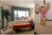 A cozy bedroom with a single bed covered in a red striped blanket, a window with green curtains, a potted plant, framed pictures on the windowsill, a chair, and a wall-mounted mirror with hats and a bag hanging on hooks nearby.