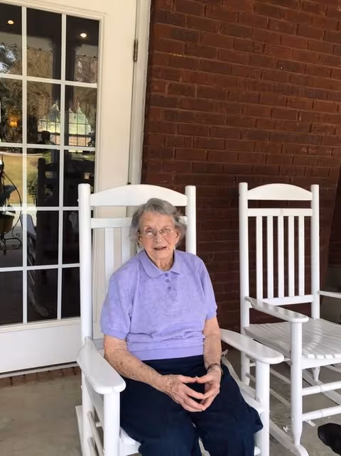 An elderly woman wearing a light purple polo shirt and dark pants is sitting on a white wooden rocking chair on a porch with a brick wall and glass door behind her. Another empty white rocking chair is next to her.