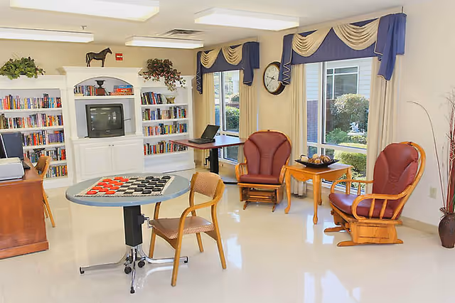 Bright senior living lounge with bookshelves, a chess table, red chairs, and large windows.