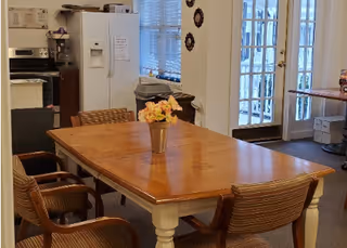 Wood dining table with a small vase of flowers and four chairs in a communal dining area next to a kitchen with a refrigerator and French doors.