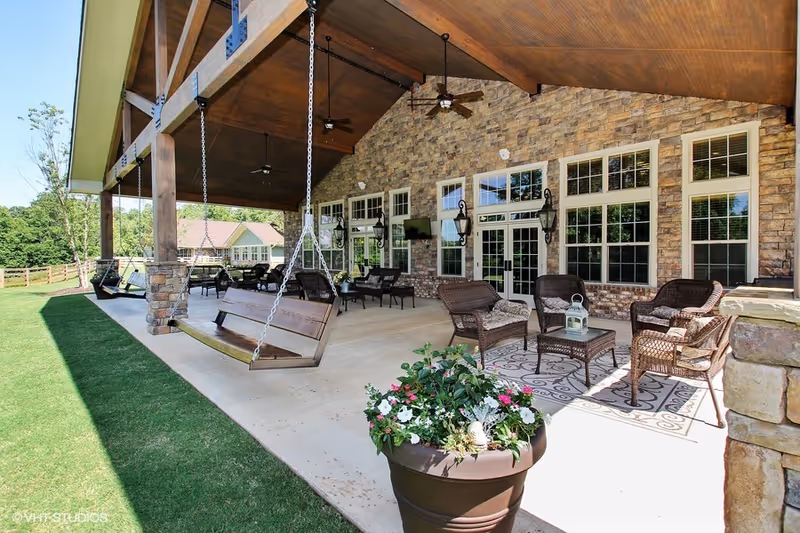 Covered outdoor patio area with wooden swings hanging from the ceiling, wicker chairs and tables arranged on a concrete floor, a large potted plant with flowers in the foreground, and a stone wall with multiple windows and doors in the background.