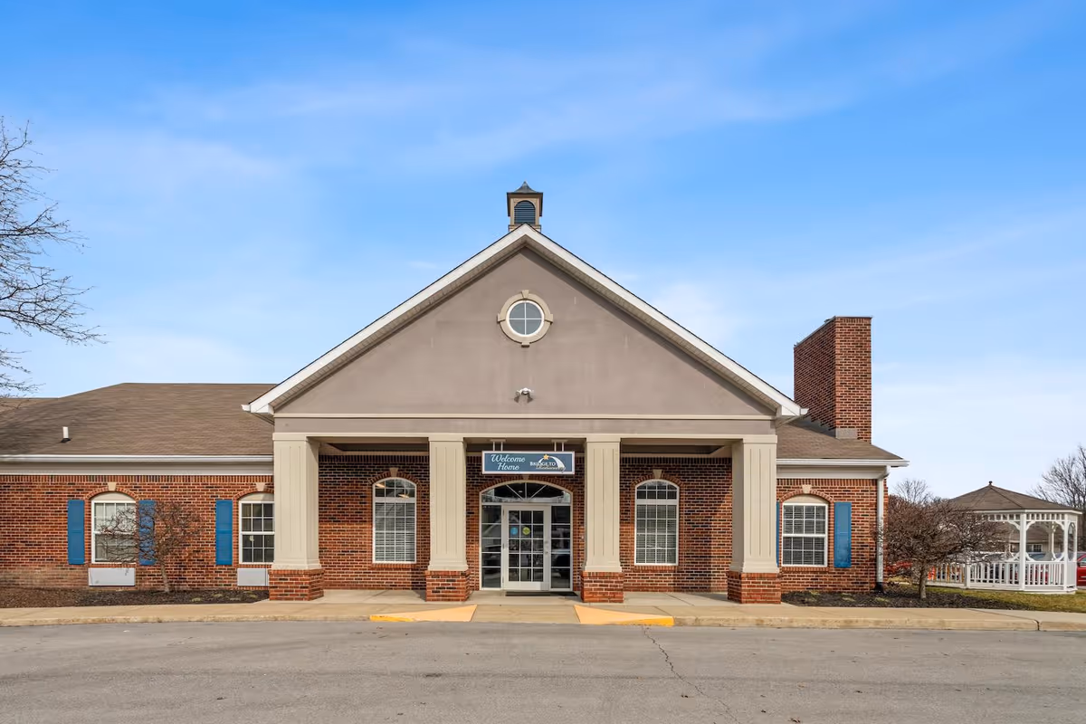 Front entrance of a brick senior living building with columns and a small white gazebo visible to the right.