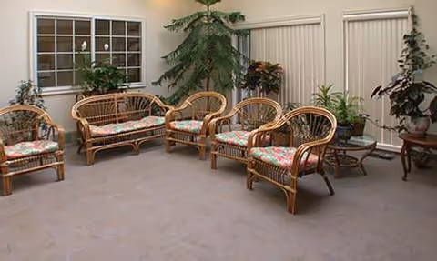 A sunroom or enclosed patio area with wicker furniture including a loveseat and four chairs with floral cushions, several potted plants, a glass-top coffee table, and windows with blinds.
