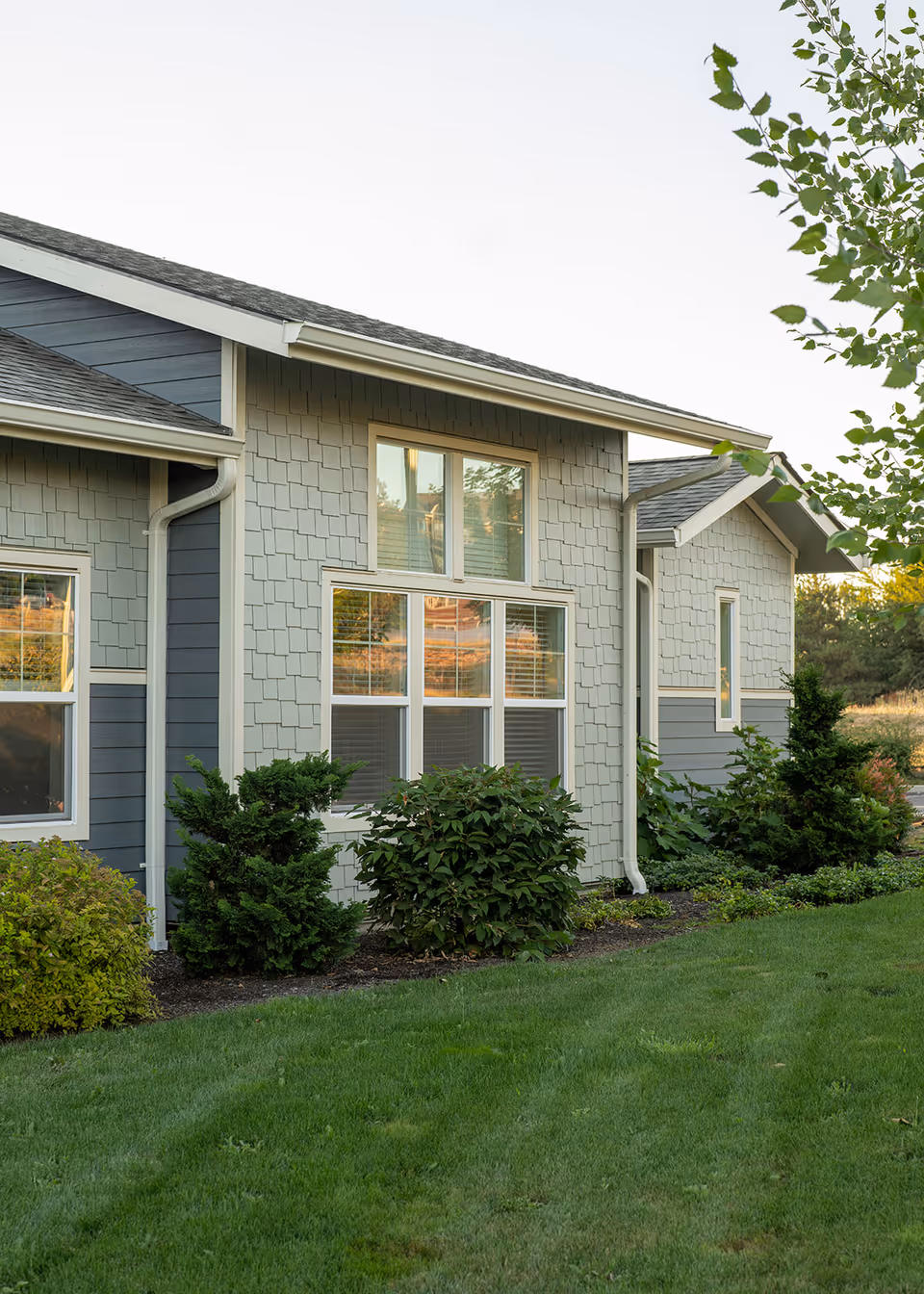 Exterior view of a single-story building with gray and blue siding, multiple windows, and a well-maintained lawn with shrubs and small trees in front.