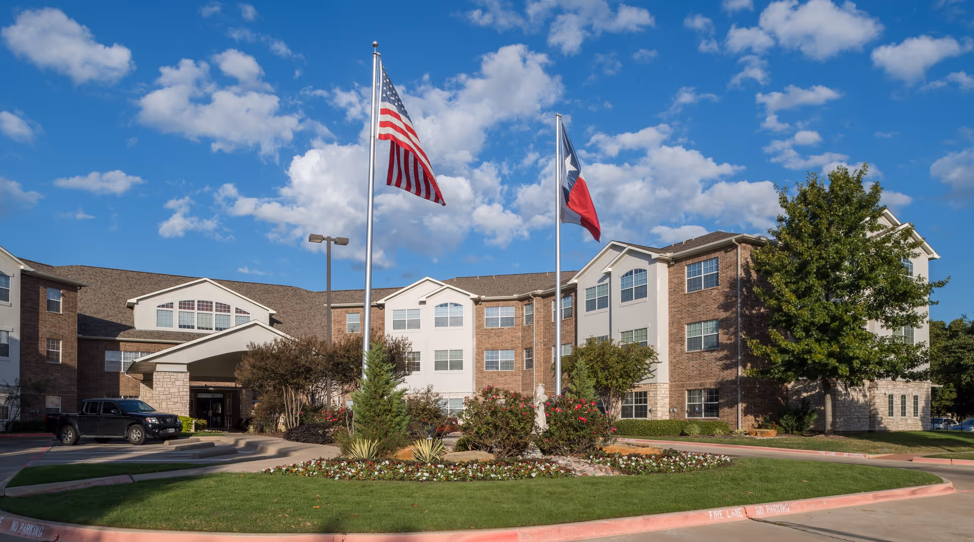 Exterior view of Mountain Creek Retirement Living facility showing a three-story building with brick and white siding, a covered entrance, two flagpoles with the American and Texas flags, landscaped greenery, and a clear blue sky with scattered clouds.