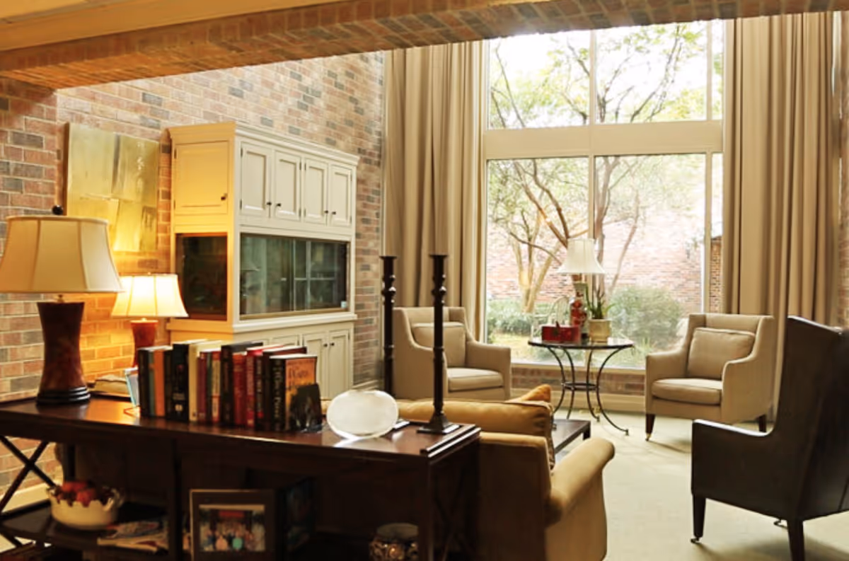 Bright living room with armchairs and a sofa arranged around a console table, a large floor-to-ceiling window overlooking a courtyard, and decorative lamps.