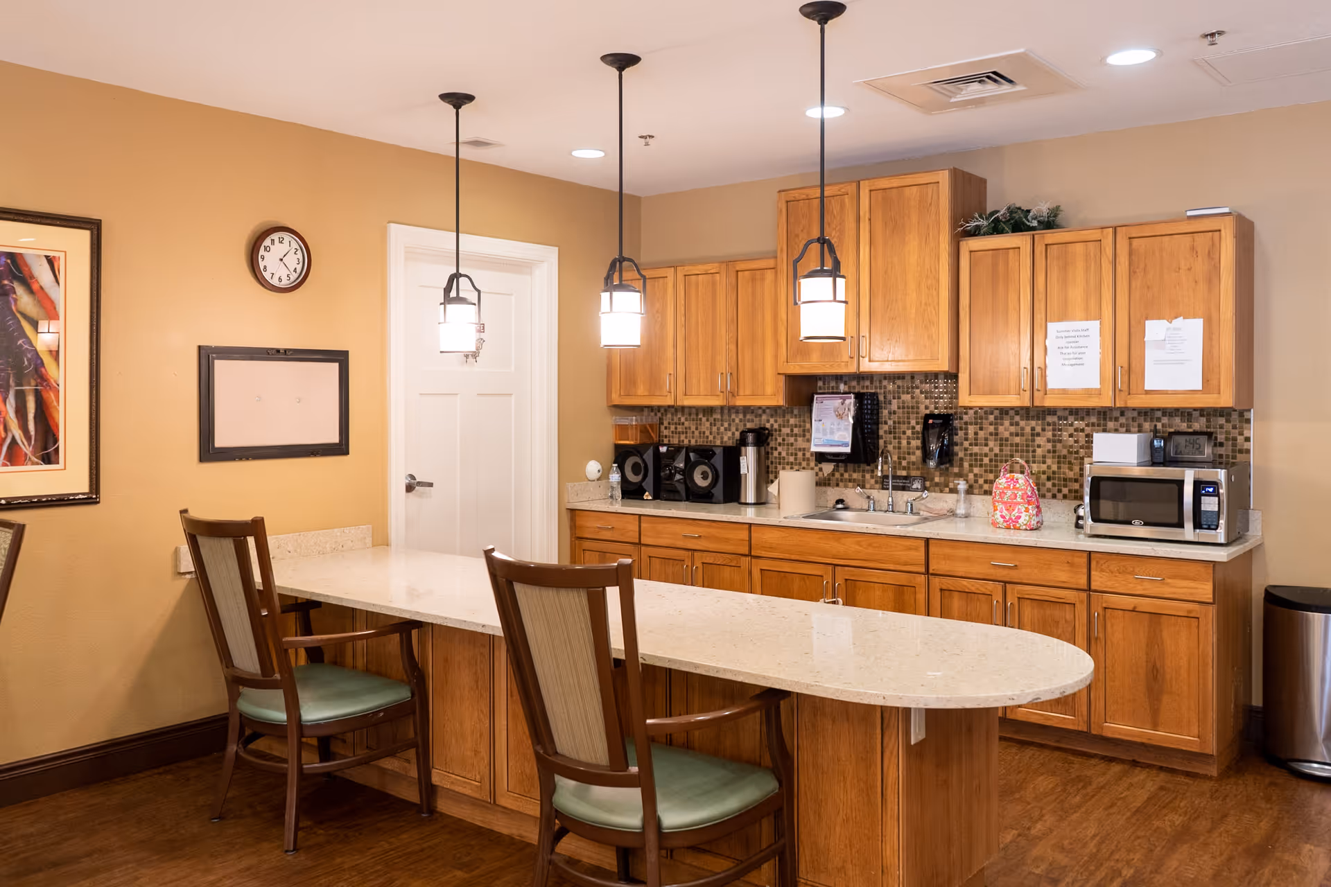 A kitchen area in an assisted living facility with wooden cabinets, a countertop island with two chairs, a microwave, a coffee maker, and pendant lights hanging from the ceiling. The walls are painted beige, and there is a clock and framed artwork on the wall.