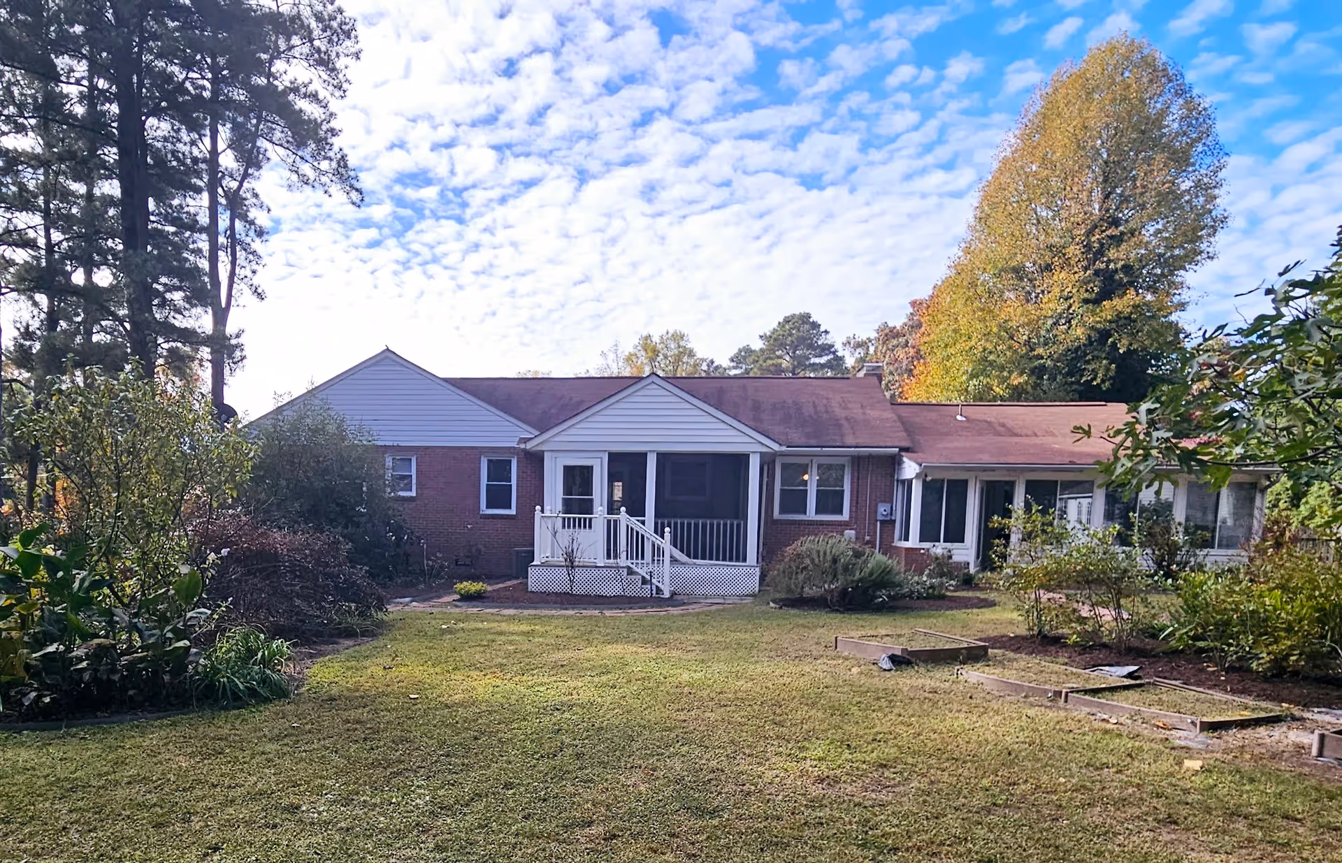 Rear exterior view of a single-story brick building with a screened porch and white railing, surrounded by a grassy yard and various trees and shrubs under a partly cloudy sky.