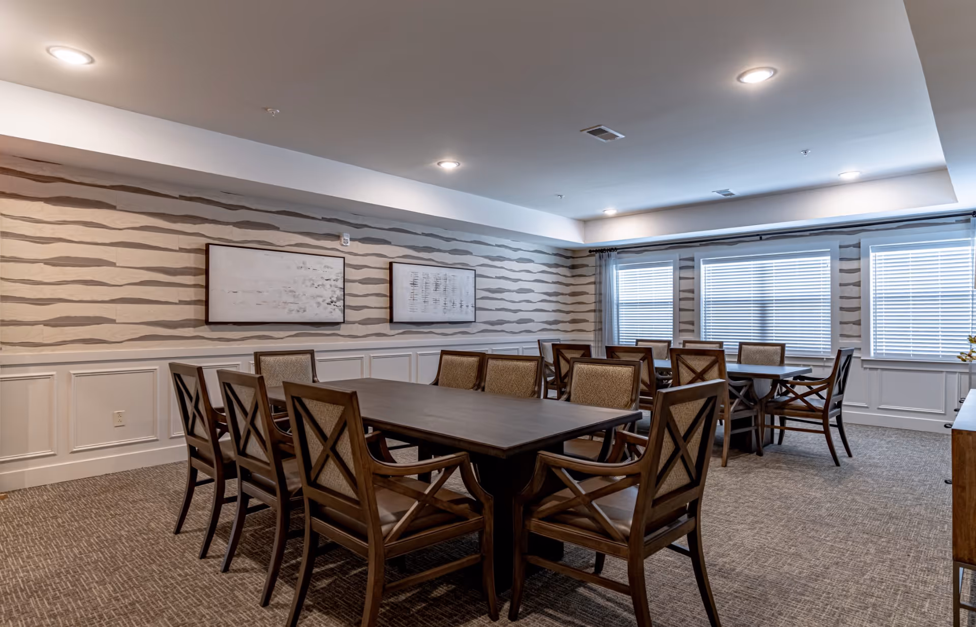 A well-lit dining room with two dark wooden tables surrounded by matching wooden chairs with cushioned seats and backs. The walls have a wavy striped pattern wallpaper above white wainscoting, and there are three large windows with white blinds letting in natural light. Two abstract paintings hang on the wall, and the ceiling has recessed lighting.