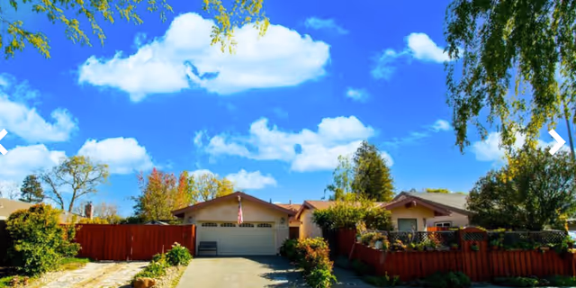 Front exterior of a single-story home with a driveway, garage, red fence, and trees under a bright blue sky.