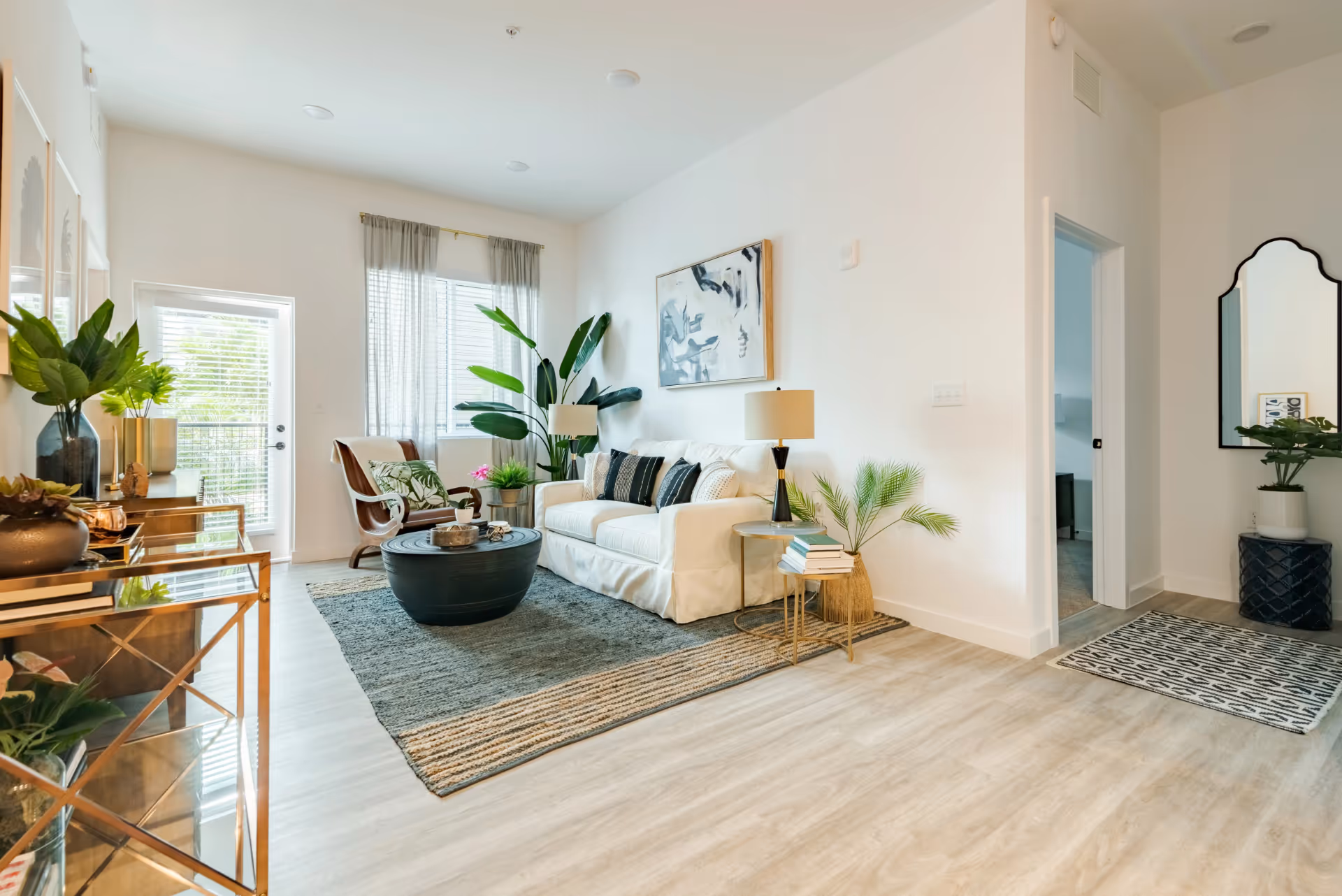 Bright and modern living room with a white sofa adorned with black and white pillows, a round black coffee table on a textured rug, a wooden armchair with a cushion, large green plants, and a side table with books and a lamp. The room has light wood flooring, white walls, a large window with sheer curtains, and a door leading outside. A mirror and additional plants are visible near an open doorway.
