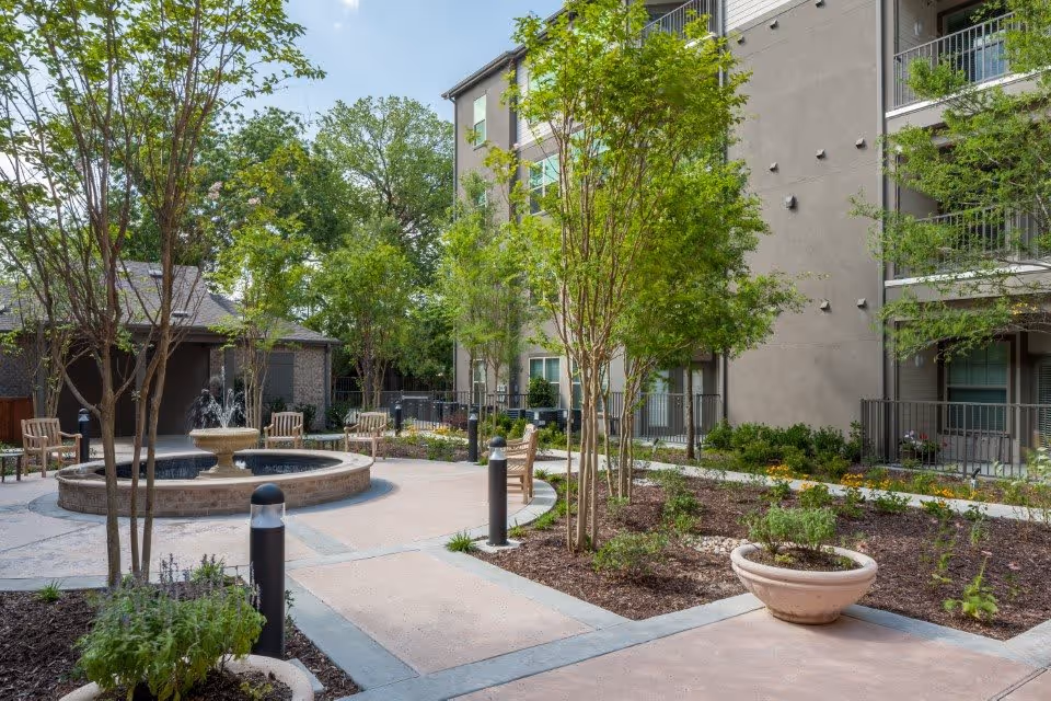 Outdoor courtyard area with a circular water fountain surrounded by benches, young trees, and landscaped garden beds. A multi-story building with balconies is visible in the background under a clear blue sky.