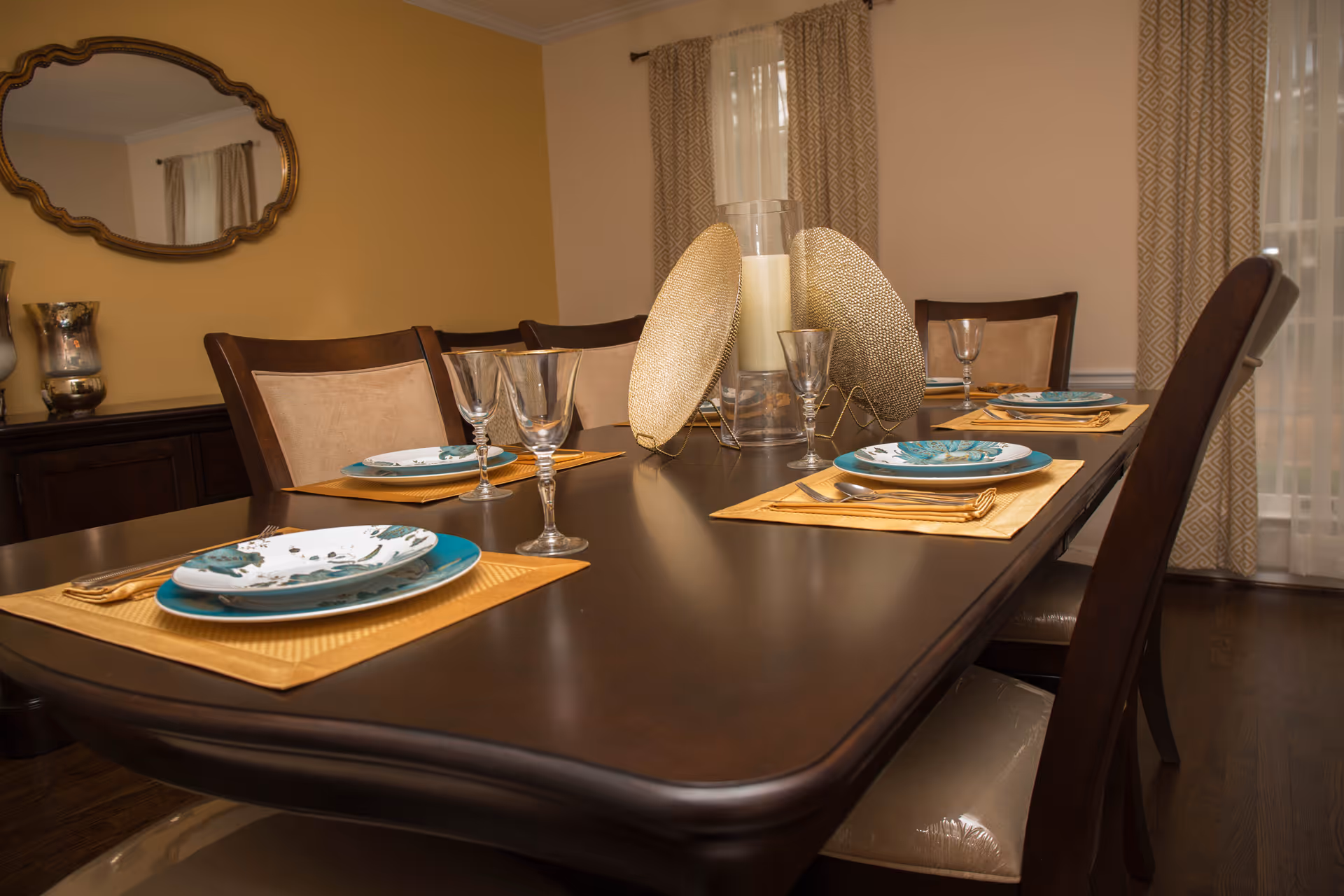 A dining room with a dark wooden table set for four people. Each place setting includes a yellow placemat, a blue and white plate, a fork, and a wine glass. The table has a centerpiece with a large candle and decorative gold plates. The room has beige walls, patterned curtains, and a large ornate mirror on one wall.