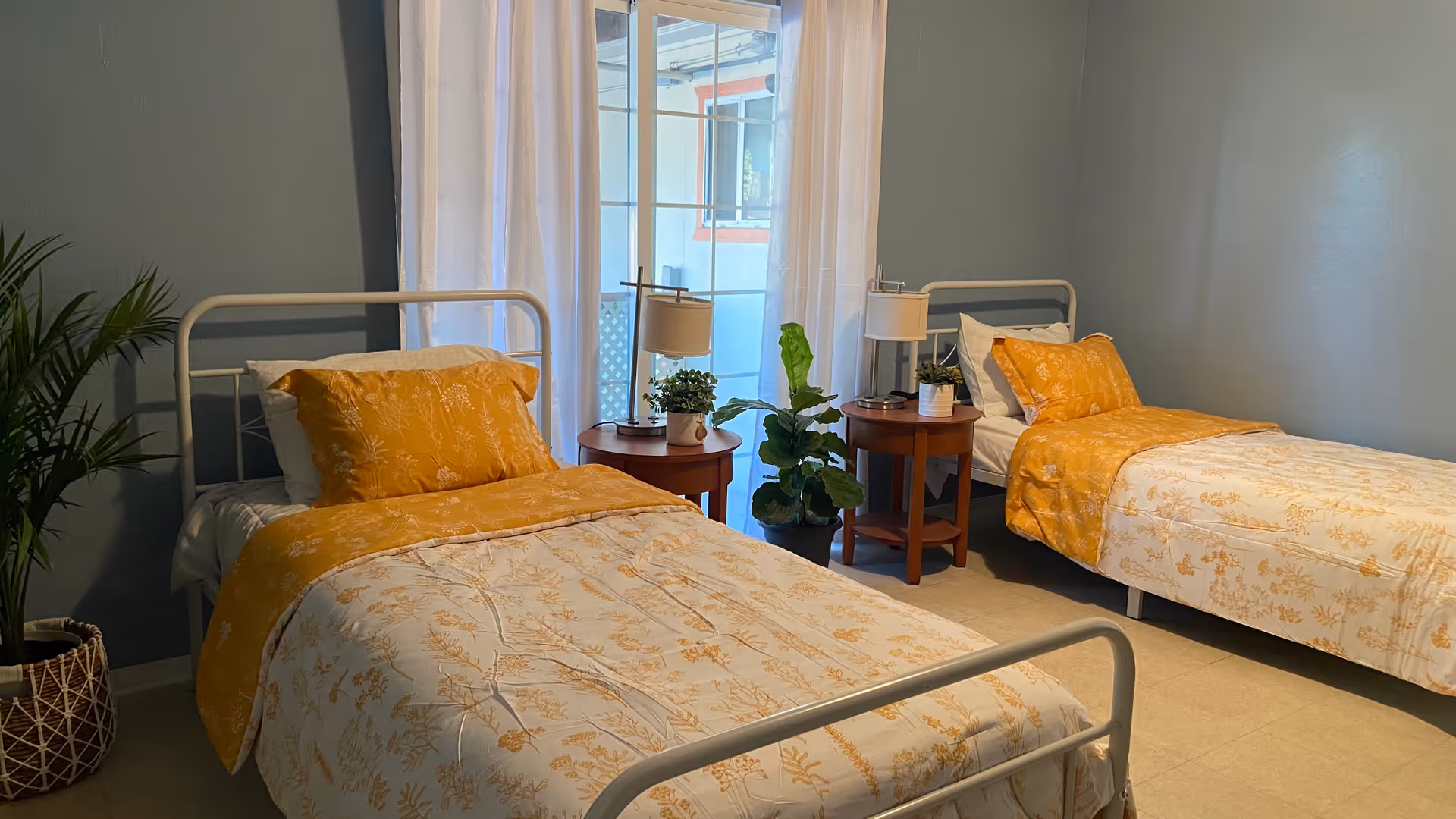 Two single beds with yellow floral bedding and white metal frames in a softly lit bedroom with bedside tables, lamps, and potted plants.