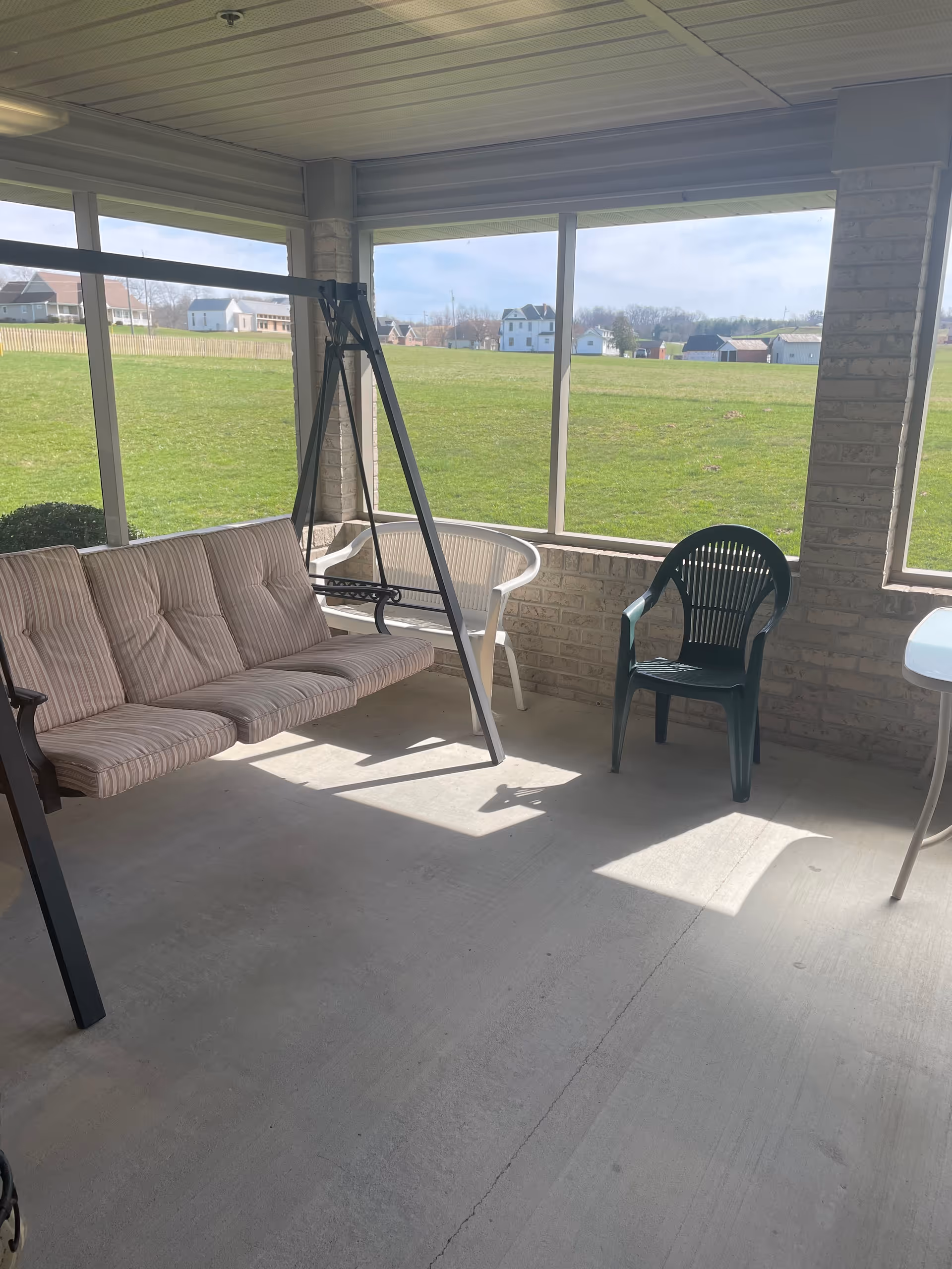 A covered patio area with a cushioned swing bench, a white plastic chair, and a green plastic chair. The patio has large screened windows overlooking a grassy field and houses in the distance.