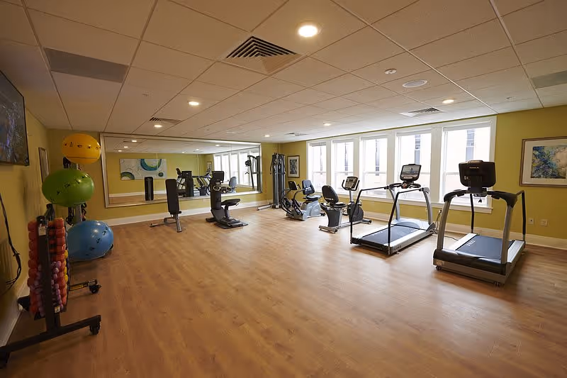 A spacious fitness room with wooden flooring and yellow walls, equipped with two treadmills, two recumbent bikes, a multi-function weight machine, and various exercise balls and dumbbells. Large windows allow natural light to fill the room, and a large mirror covers one wall.