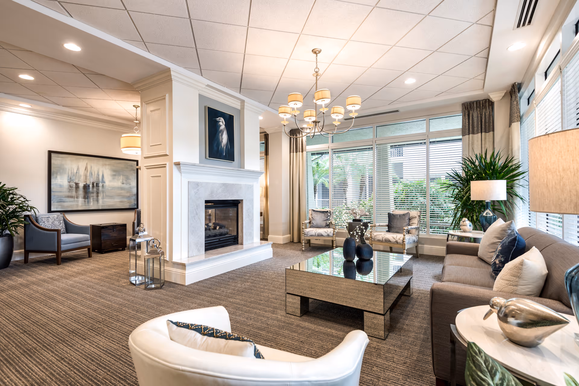 Bright communal living room with sofas, armchairs, a central mirrored coffee table, fireplace, chandelier, and large windows overlooking greenery.