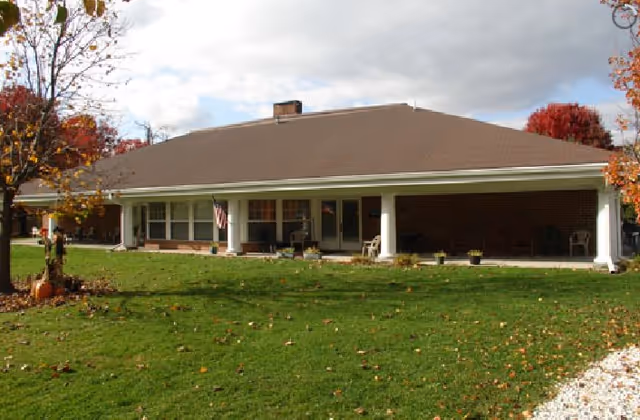 Single-story brick building with a large covered porch supported by white columns, surrounded by a green lawn and trees with autumn foliage under a partly cloudy sky.