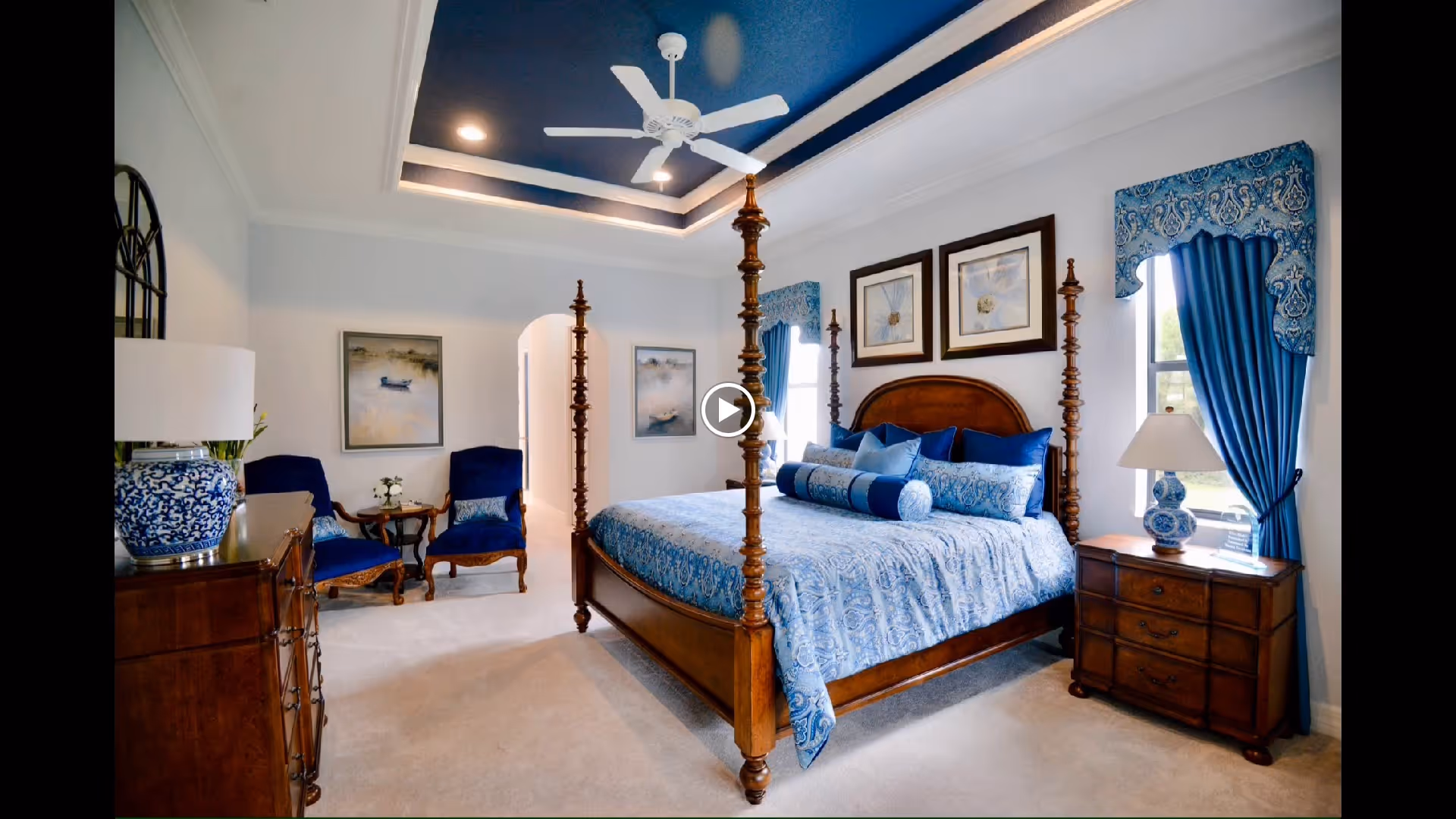 Elegant traditional bedroom with a four-poster wooden bed dressed in blue patterned bedding, matching curtains, two armchairs, bedside tables and a tray ceiling with fan.