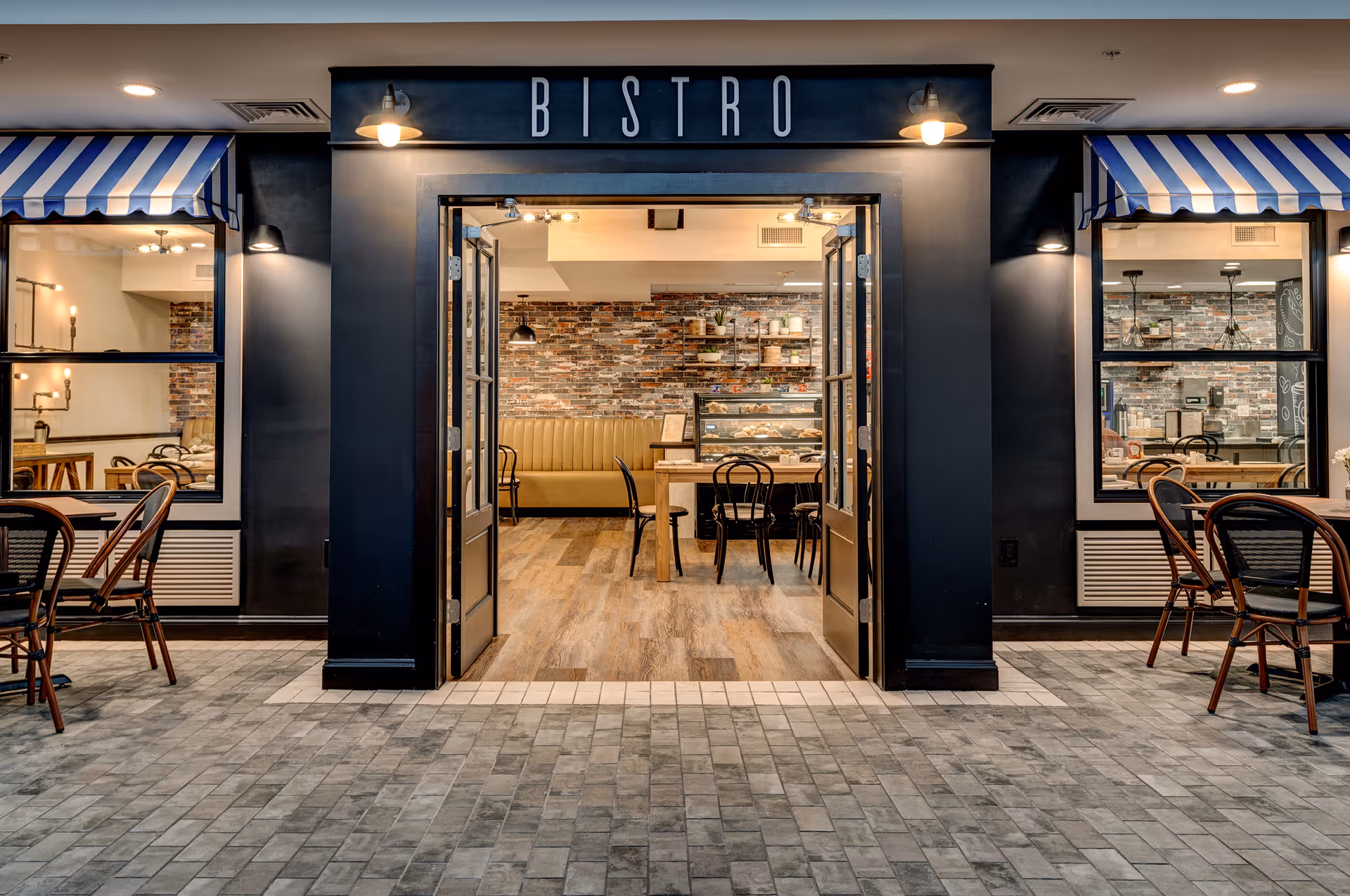 Entrance to a bistro with open double doors, showing an interior with wooden tables, chairs, and a cushioned bench against a brick wall. The exterior has black walls with blue and white striped awnings over the windows and outdoor seating with tables and chairs on a tiled floor.