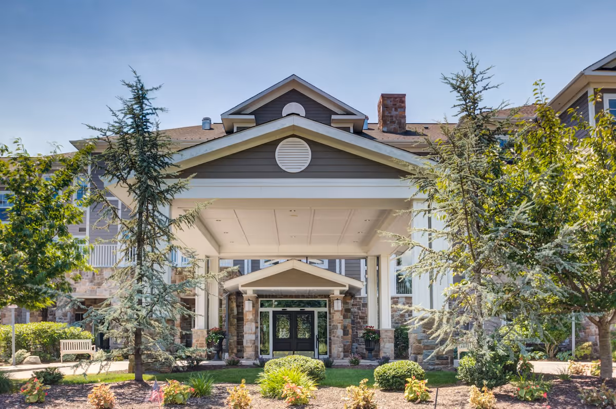 Front exterior view of a senior living facility with a covered entrance, stone and wood siding, surrounded by trees and landscaped bushes under a clear blue sky.
