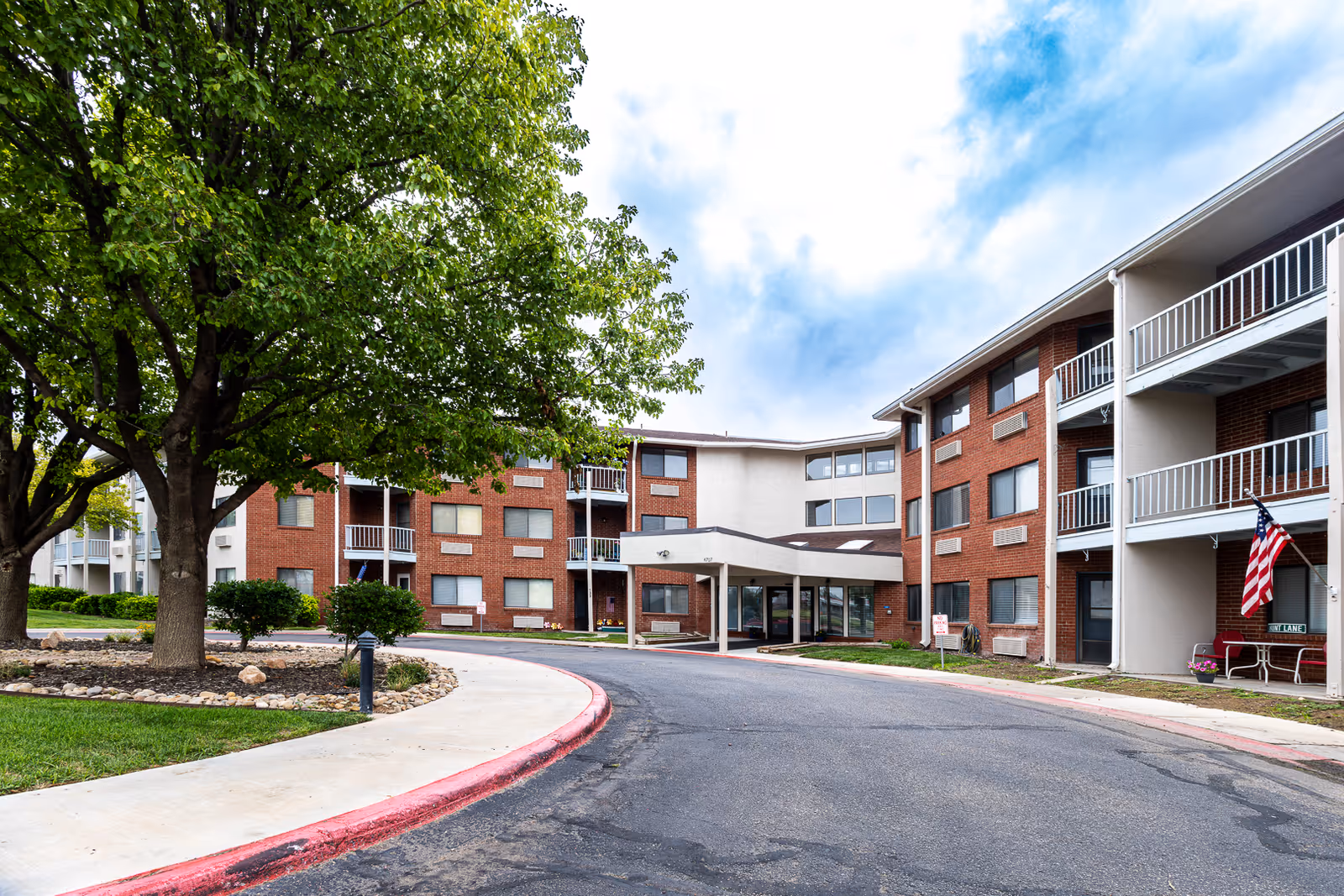 Front exterior of a three-story brick senior living building with a covered entrance, balconies, trees, and an American flag.