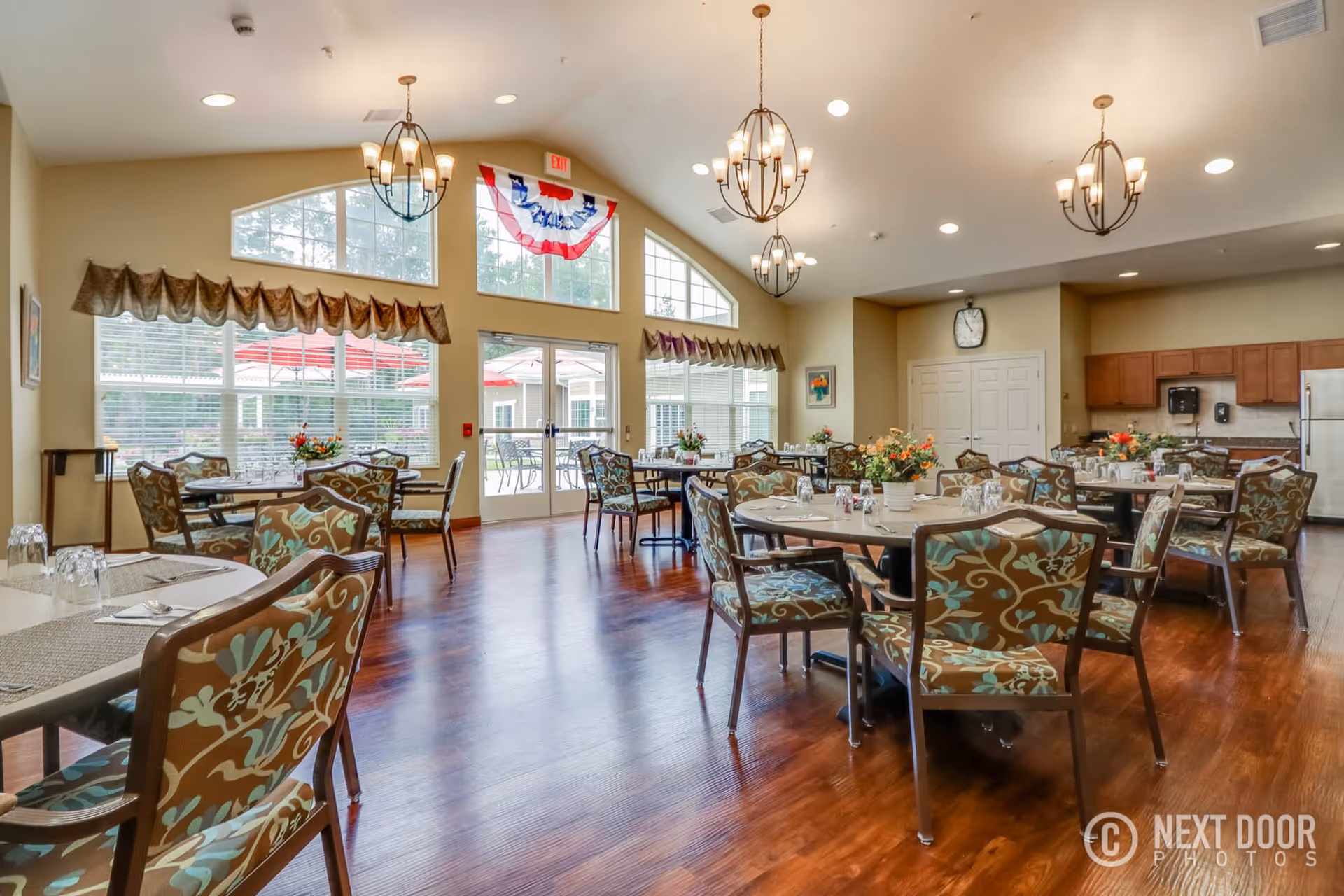 A bright and spacious dining room with multiple round tables and floral-patterned chairs. The room features large windows with valances, allowing natural light to fill the space. There are chandeliers hanging from the ceiling, and a kitchen area with wooden cabinets and a refrigerator is visible in the background. The floor is made of polished wood, and there are floral centerpieces on the tables. Outside the windows, red umbrellas and patio furniture can be seen.