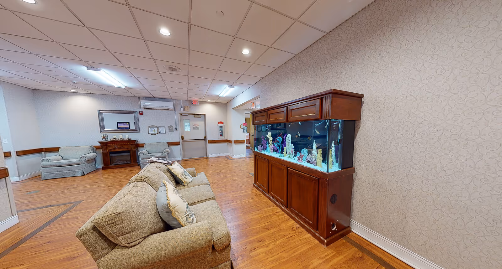 A spacious living room area with wooden flooring, beige walls, and a ceiling with recessed lighting. The room features a large wooden aquarium filled with colorful coral decorations on the right side, a beige sofa with cushions in the foreground, and two armchairs near a fireplace with a mirror above it in the background. There is a door and some wall-mounted items visible at the far end of the room.