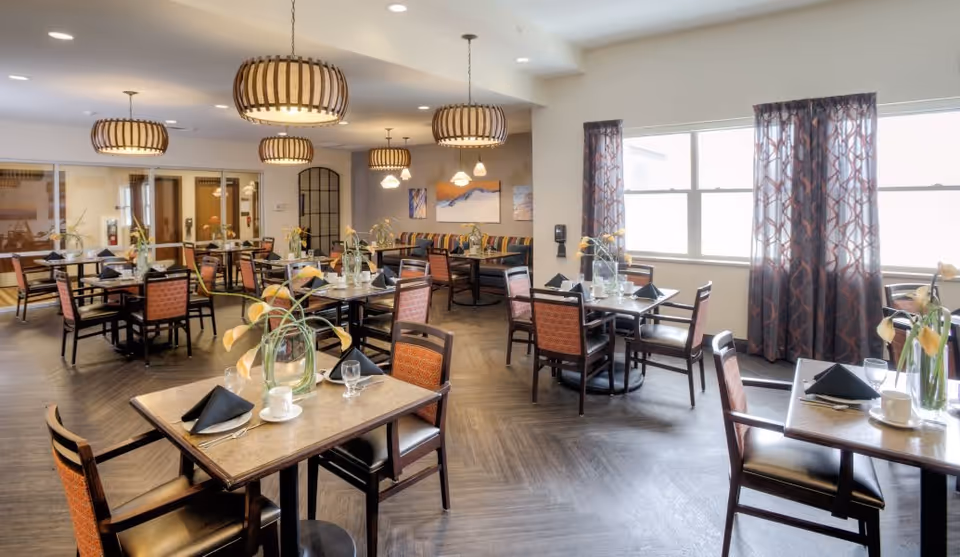 Dining room with multiple set tables and chairs, pendant lighting, and large windows in a senior living facility.
