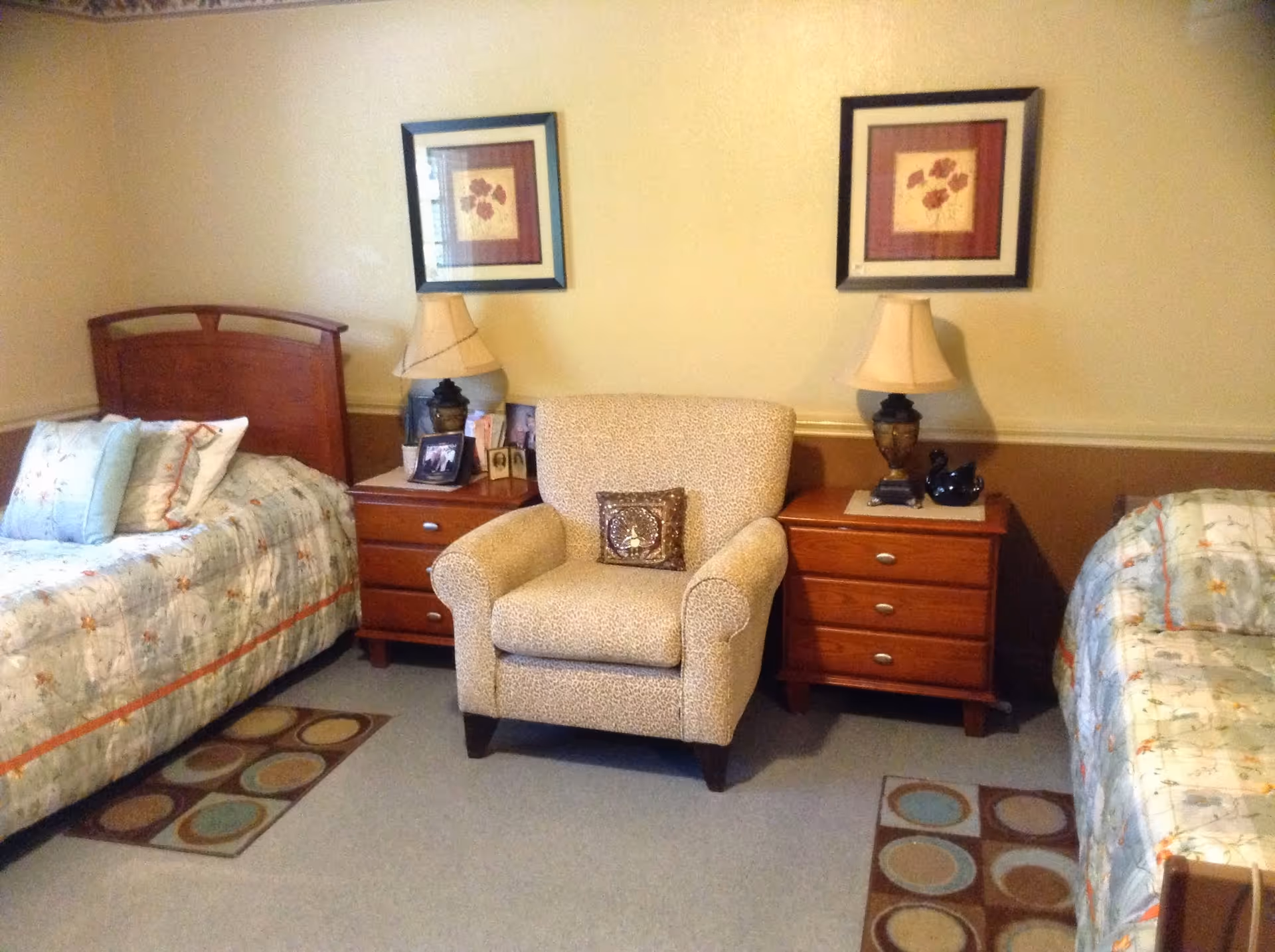 A cozy bedroom with two single beds on either side of a beige armchair. Each bed has floral patterned bedding and pillows. Between the beds are two wooden nightstands with table lamps, framed photos, and decorative items. Above each nightstand hangs a framed floral artwork. The room has a warm, inviting atmosphere.