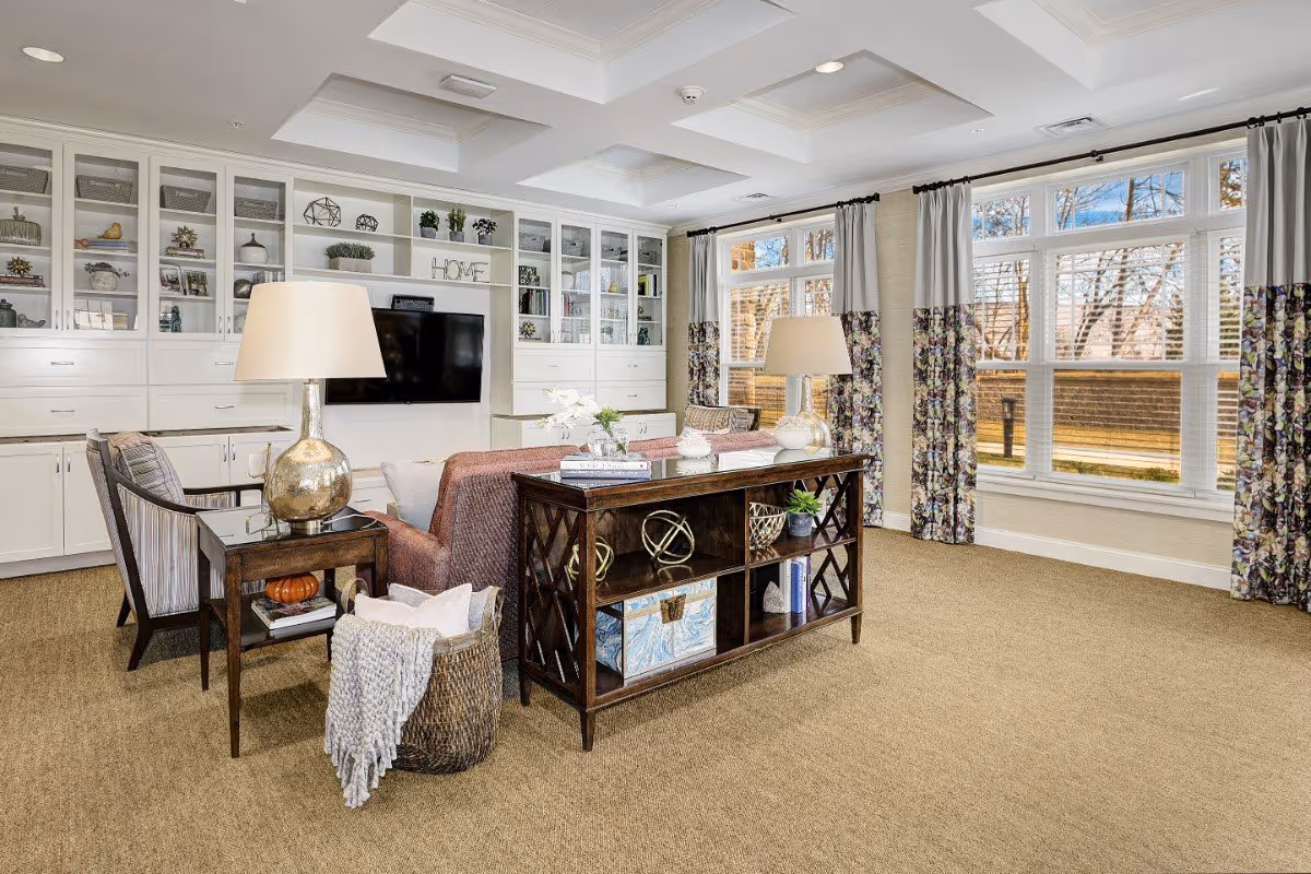 A bright and spacious living room with large windows covered by floral curtains, beige carpet, and a coffered ceiling. The room features a TV mounted on a white built-in cabinet wall with shelves displaying decorative items. There is a sofa with a wooden console table behind it, two armchairs, and side tables with lamps. A basket with a blanket and pillows is placed beside the sofa.