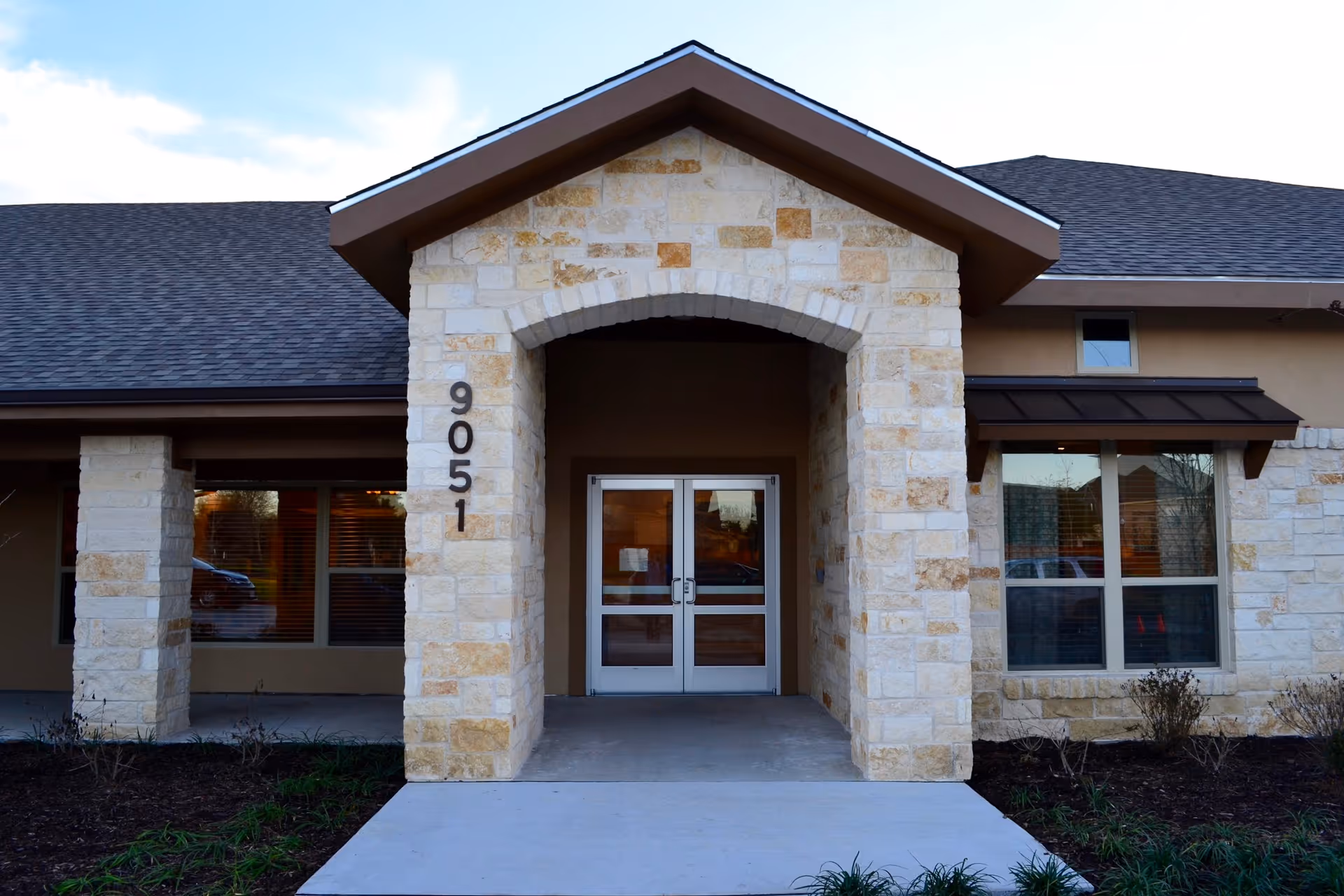 Stone-faced front entrance of a single-story facility with double glass doors and the number 9051 on a column.