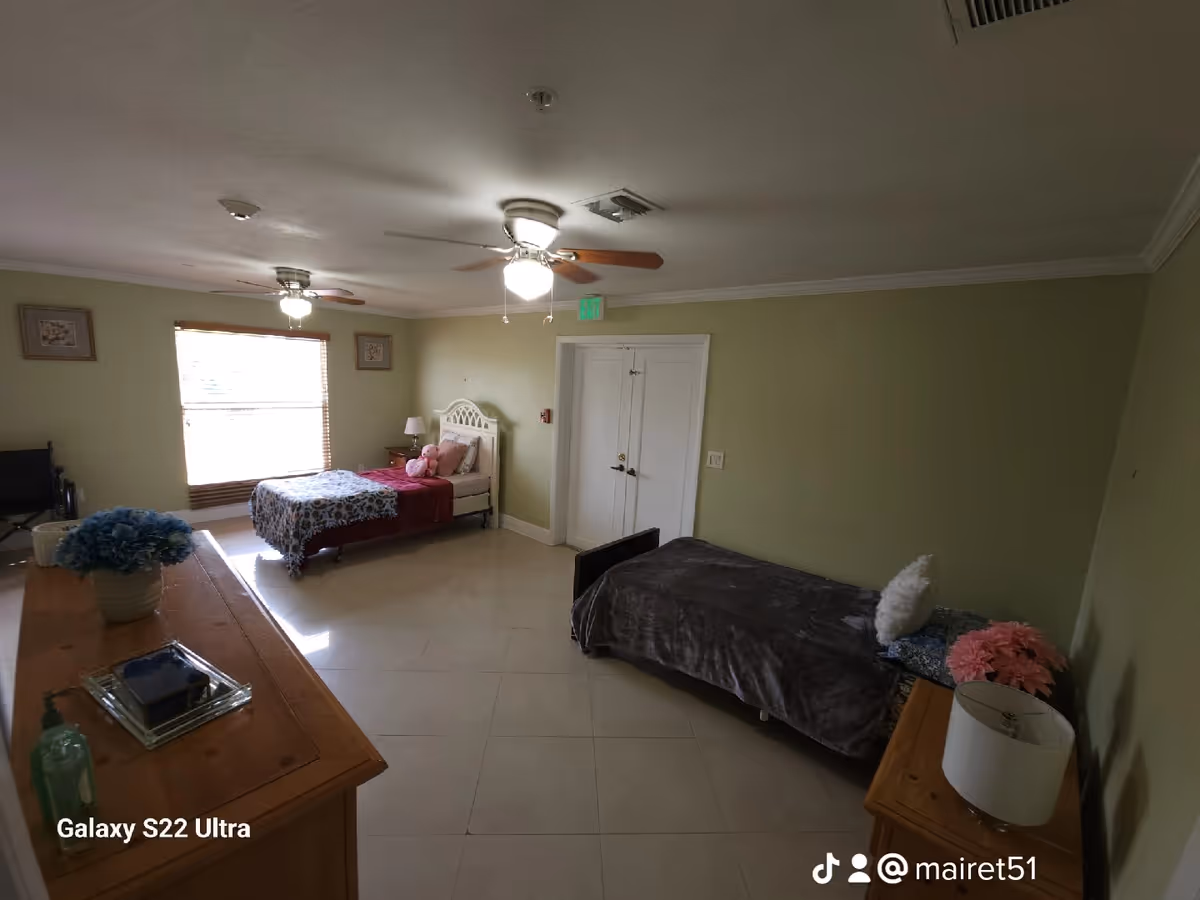A bedroom in an assisted living facility with two single beds, one near a window with floral bedding and a teddy bear, and the other with a dark blanket and pillows. The room has light green walls, a ceiling fan with lights, a wooden dresser with a vase of blue flowers, and a small lamp on a bedside table.