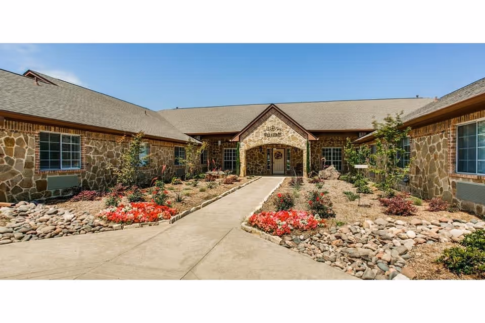 Front exterior view of Avalon Memory Care facility with stone walls, a central entrance with an archway, landscaped garden beds with flowers and rocks, and a clear blue sky.
