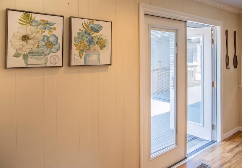 Interior view of a room with light-colored walls featuring two framed floral paintings. There is a glass door with blinds partially open leading to an outdoor deck area. On the wall next to the door, there are decorative wooden utensils hanging.